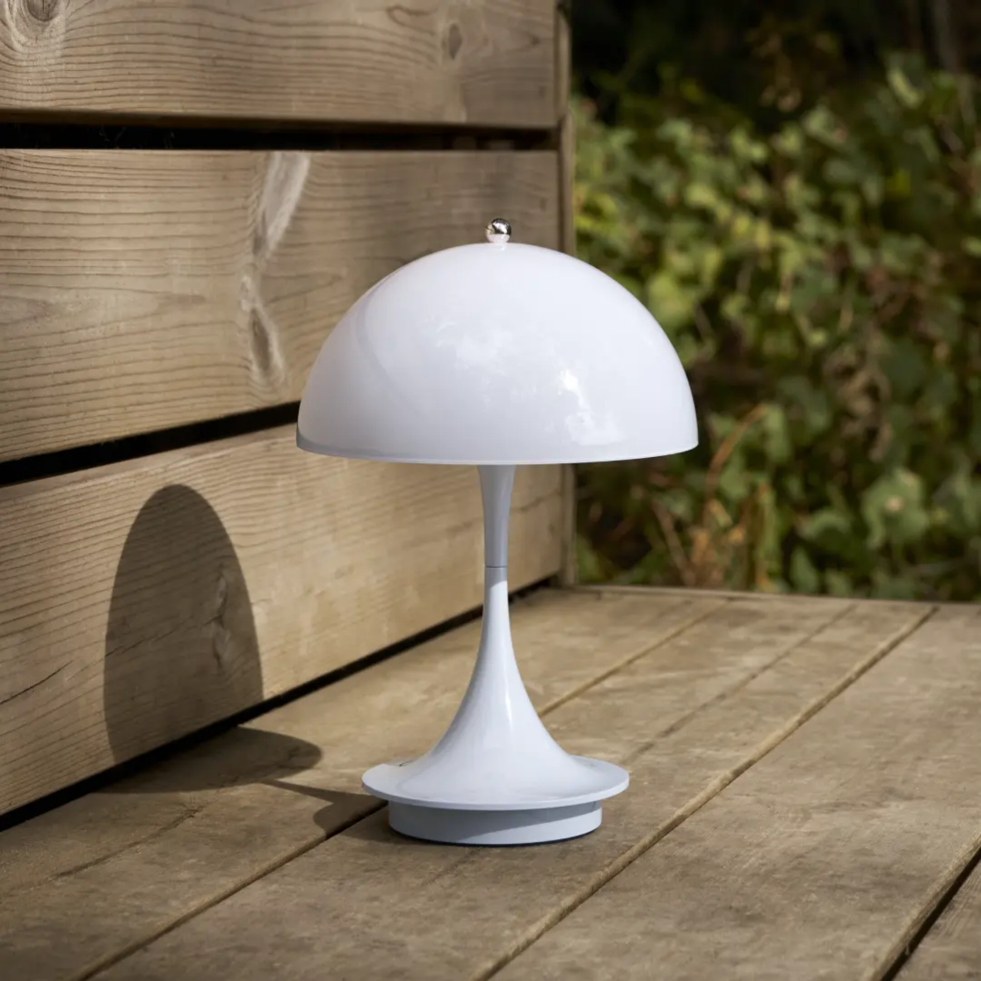 The pale blue opal Panthella 160 Portable Lamp from Louis Poulsen in a garden on top of a wooden bench.