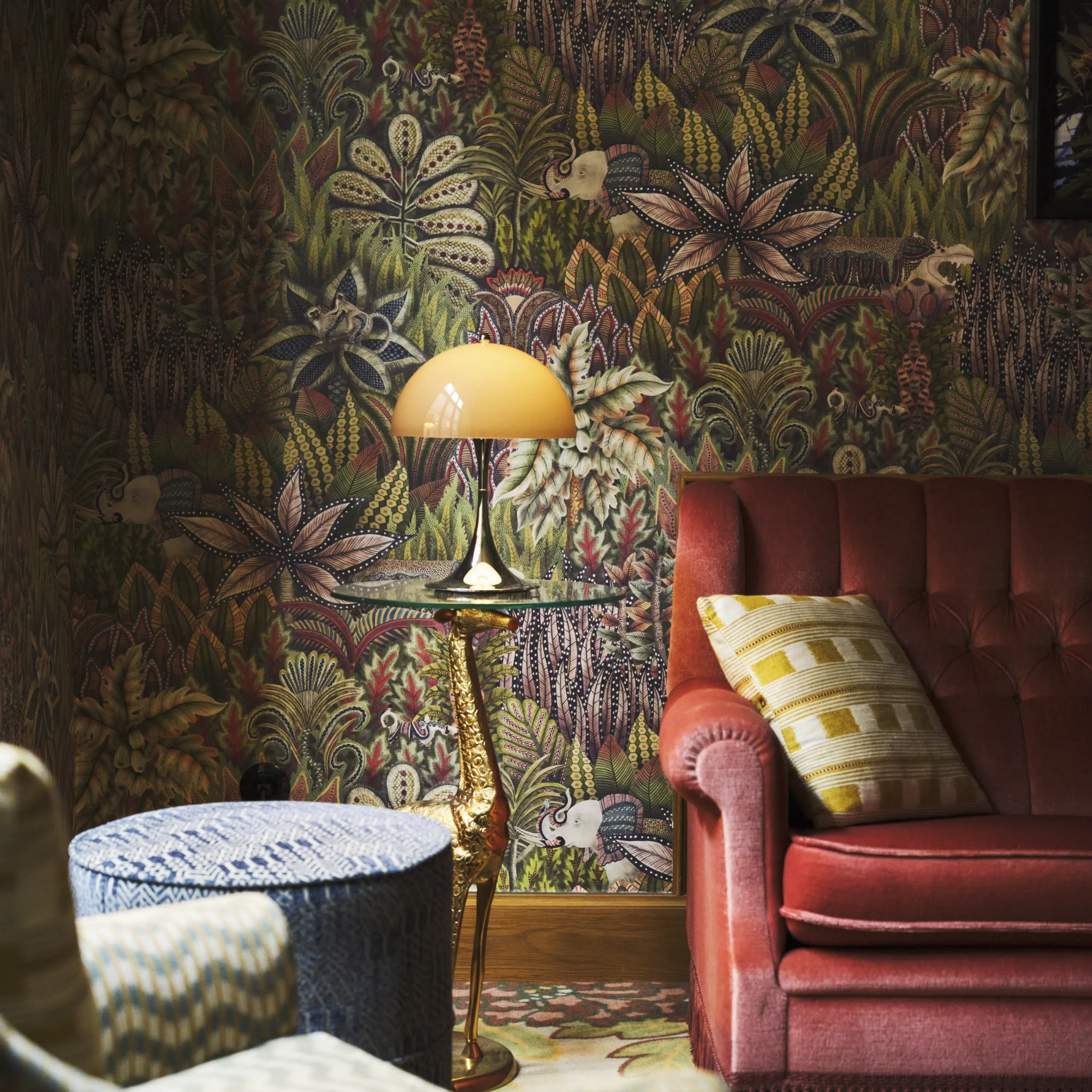 The 13.2 inch brown Panthella Portable Lamp: The Originals from Louis Poulsen in a lounge with a jungle inspired wallpaper, a sofa with cushions, a pouf, and a lounge chair.