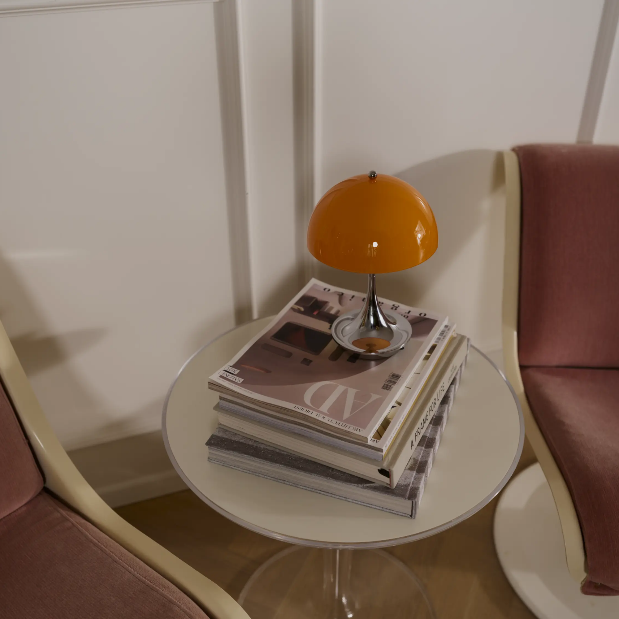 The 9.5 inch orange Panthella Portable Lamp: The Originals from Louis Poulsen on top of magazine in a living space, beside two chairs.