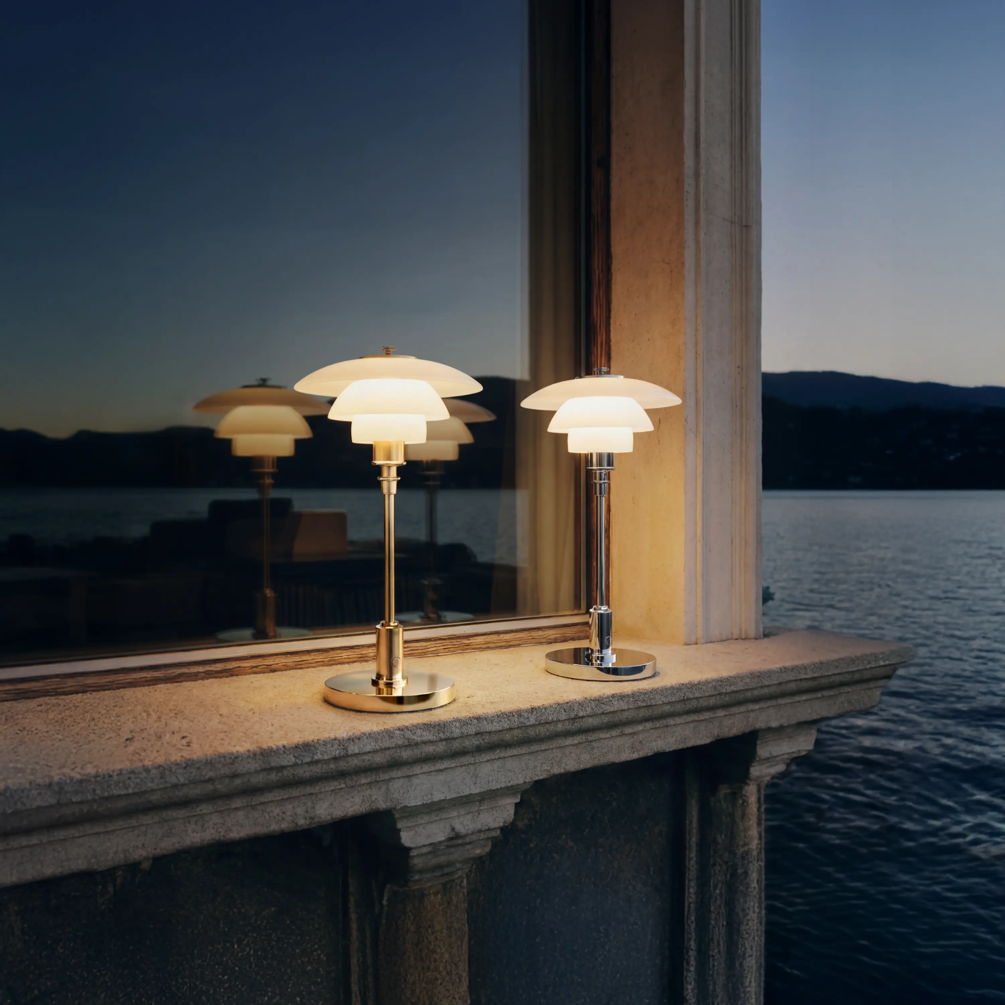 The chrome PH 1/1 Portable Lamp from Louis Poulsen in a living room beside a lake with mountains in the background.
