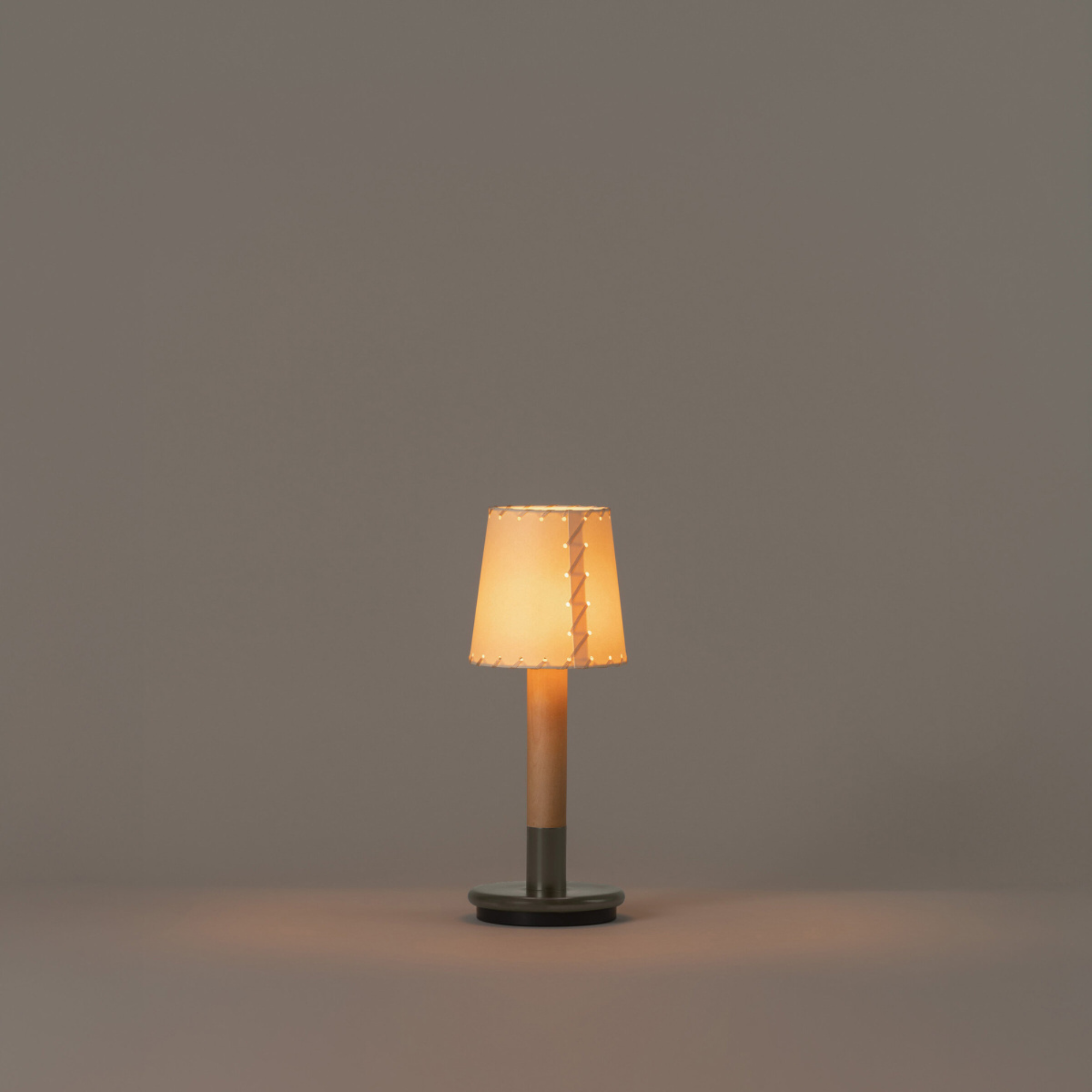 The Básica Mínima Batería Cordless Lamp from Santa & Cole with the stitched beige parchment shade turned on in a studio.