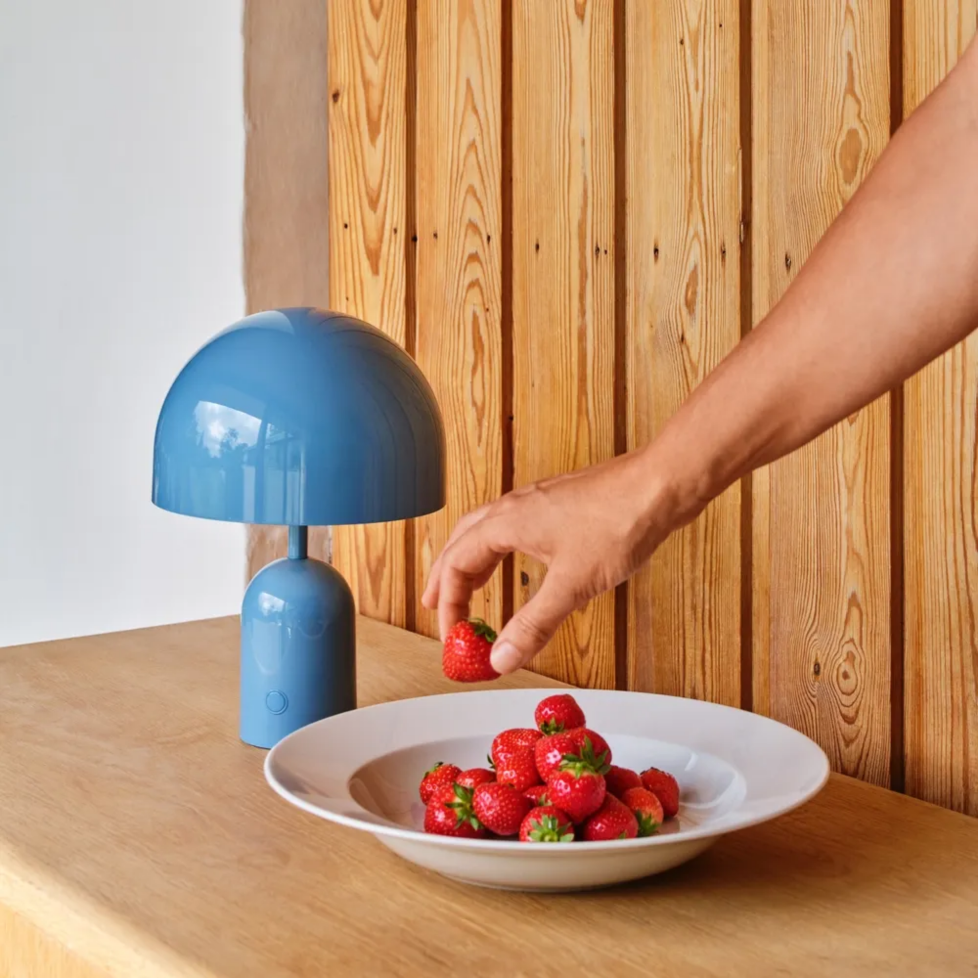 The baby blue Bell Cordless Lamp from Tom Dixon in a living space on a wooden counter.