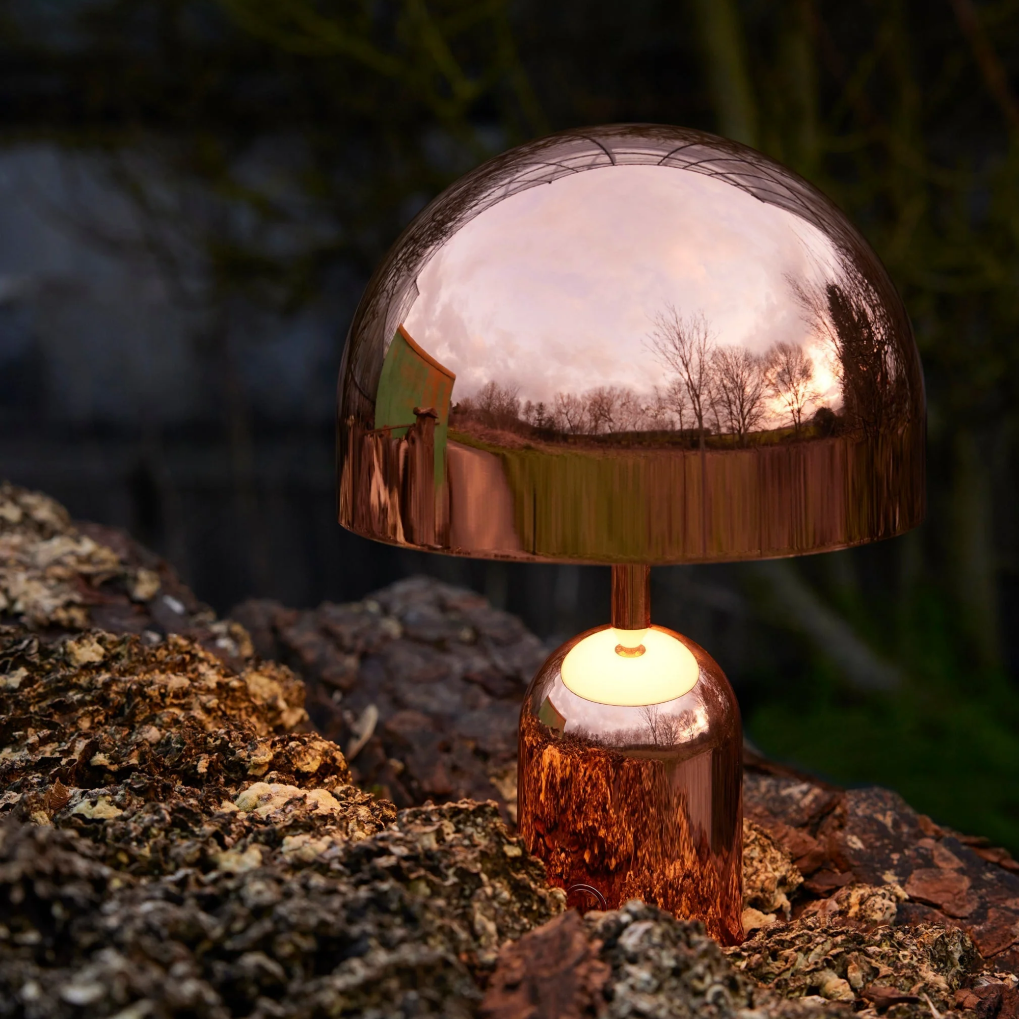 The copper Bell Cordless Lamp from Tom Dixon in a garden with trees in the background.