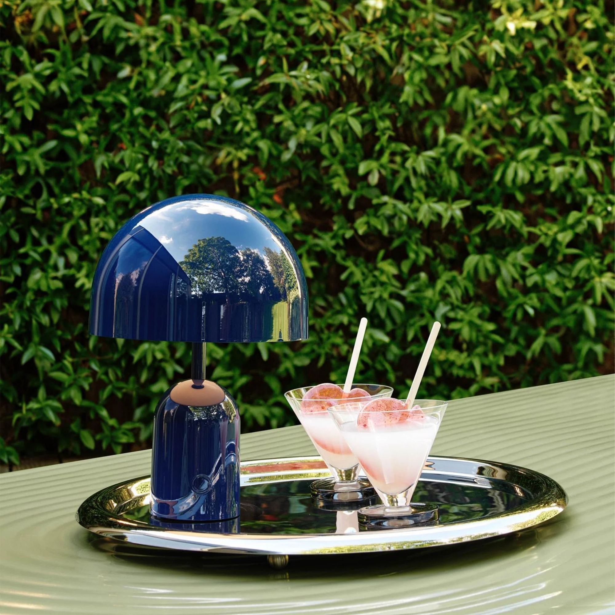 The indigo Bell Cordless Lamp from Tom Dixon in a garden beside a pair of drinks.