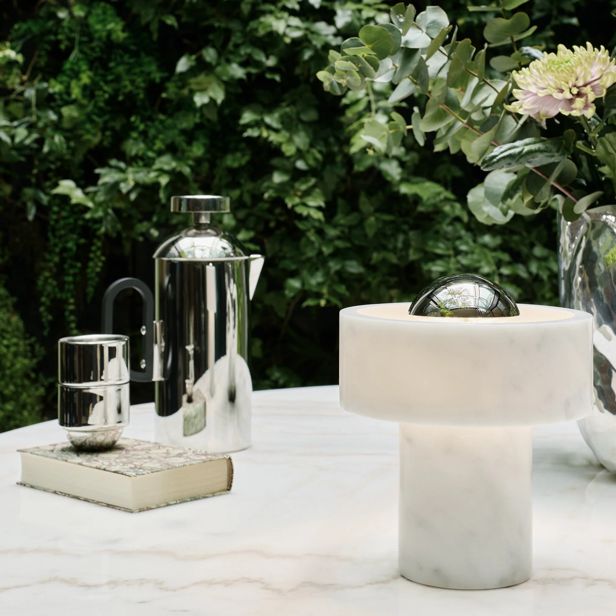 The silver Stone Cordless Lamp from Tom Dixon in a garden on top of a marble table.