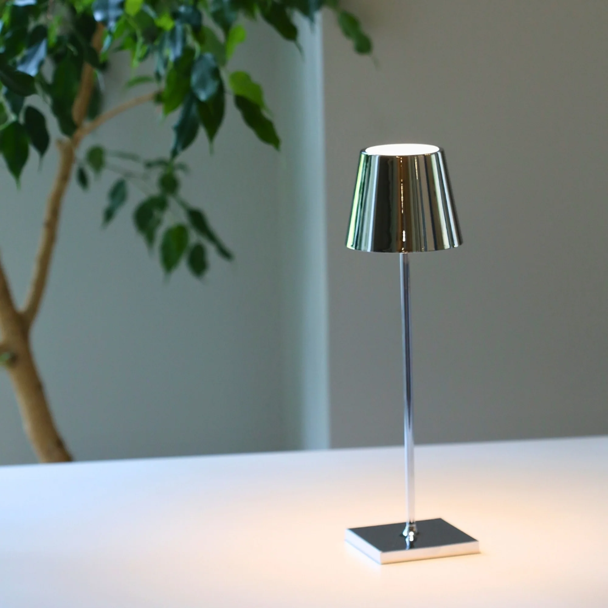 The glossy chrome Poldina Pro from Zafferano in a home office on top of a table near a tree.