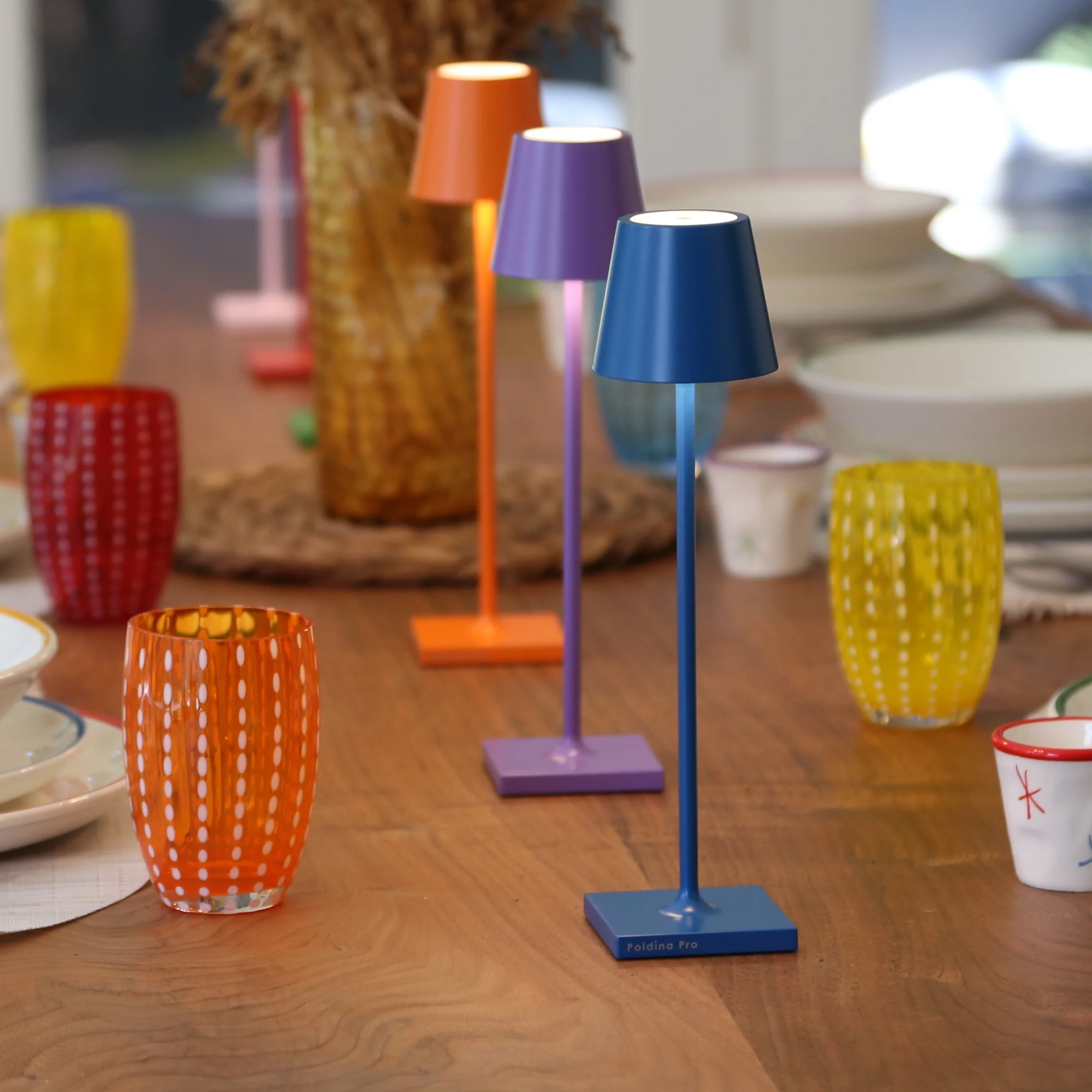 The capri blue Poldina Pro Micro Cordless Lamp from Zafferano on a table at an outdoor dinner party with various other colors.