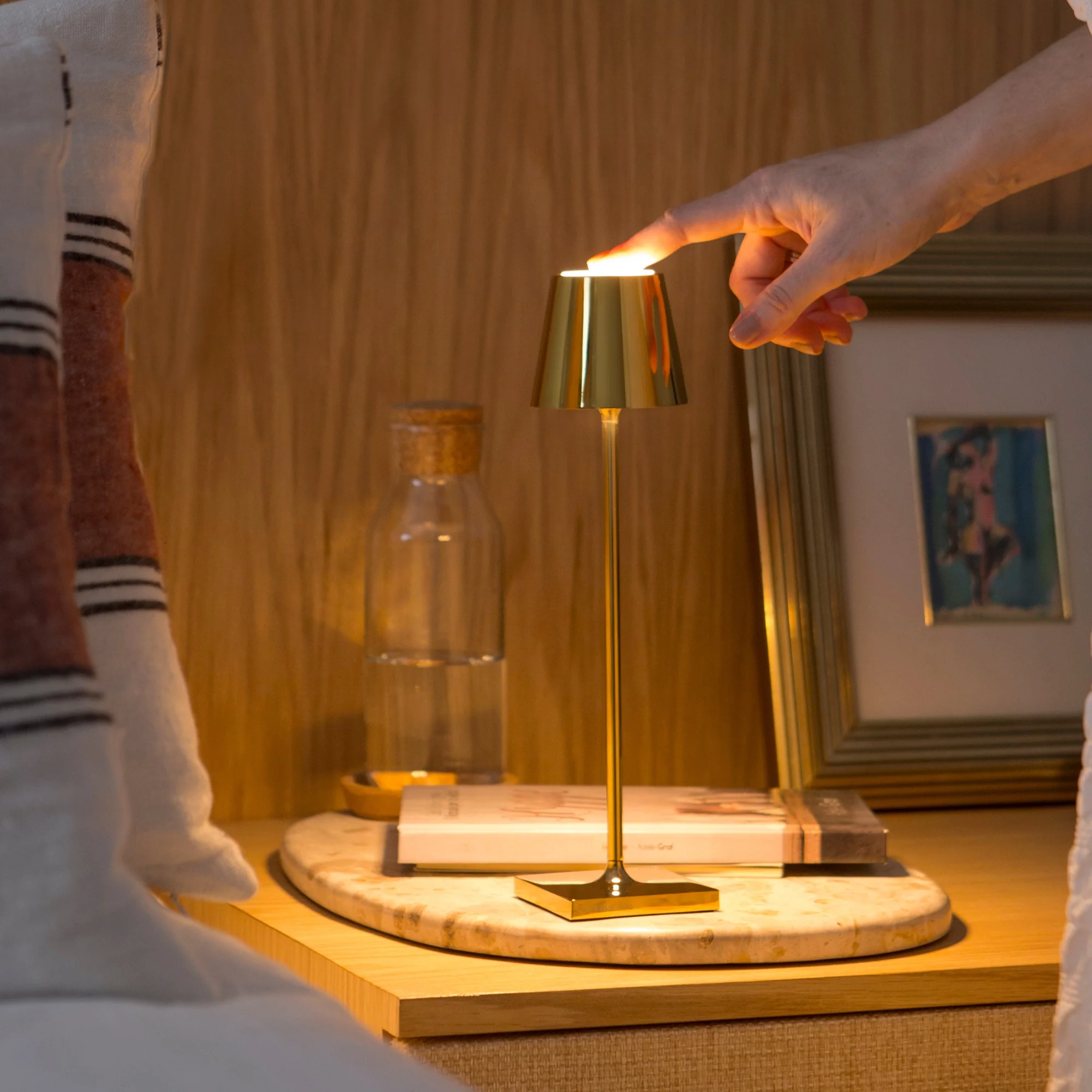 The glossy gold Poldina Pro Micro Cordless Lamp from Zafferano in a bedroom, on a bedside table being used.