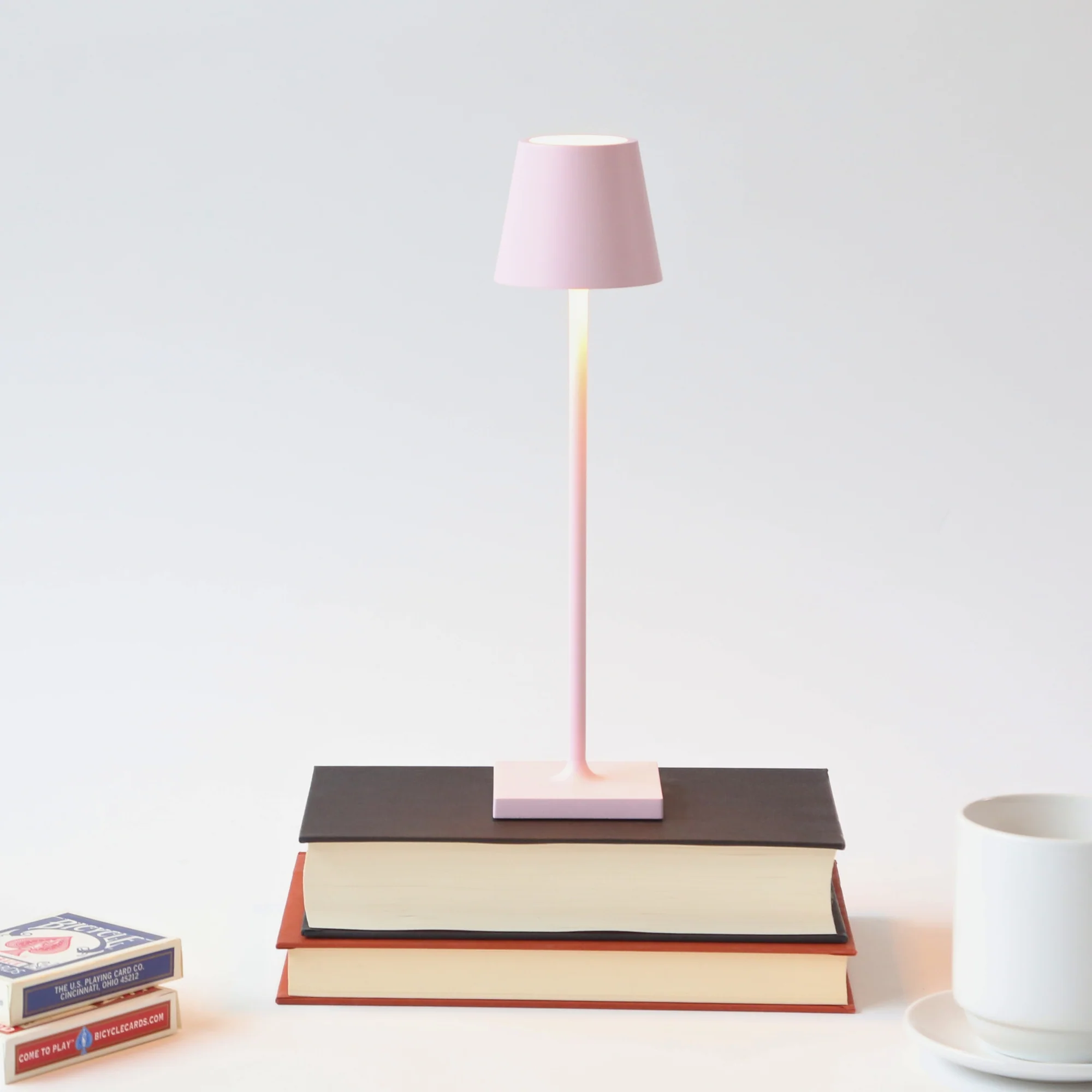 The pink Poldina Pro Micro Cordless Lamp from Zafferano in a library studio on top of a stack of books.