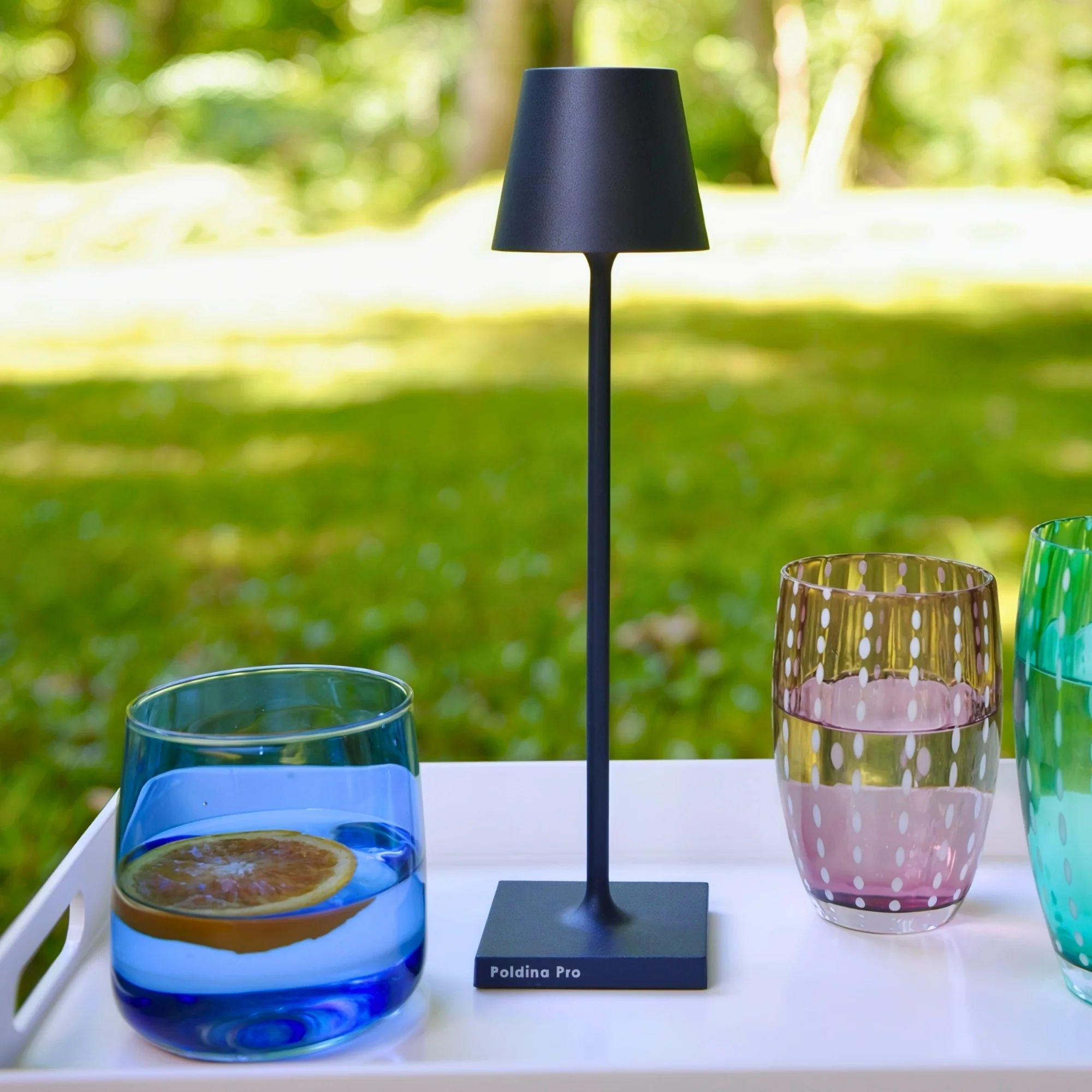 The steel blue Poldina Pro Micro Cordless Lamp from Zafferano in a garden on top of a tray with drinks.