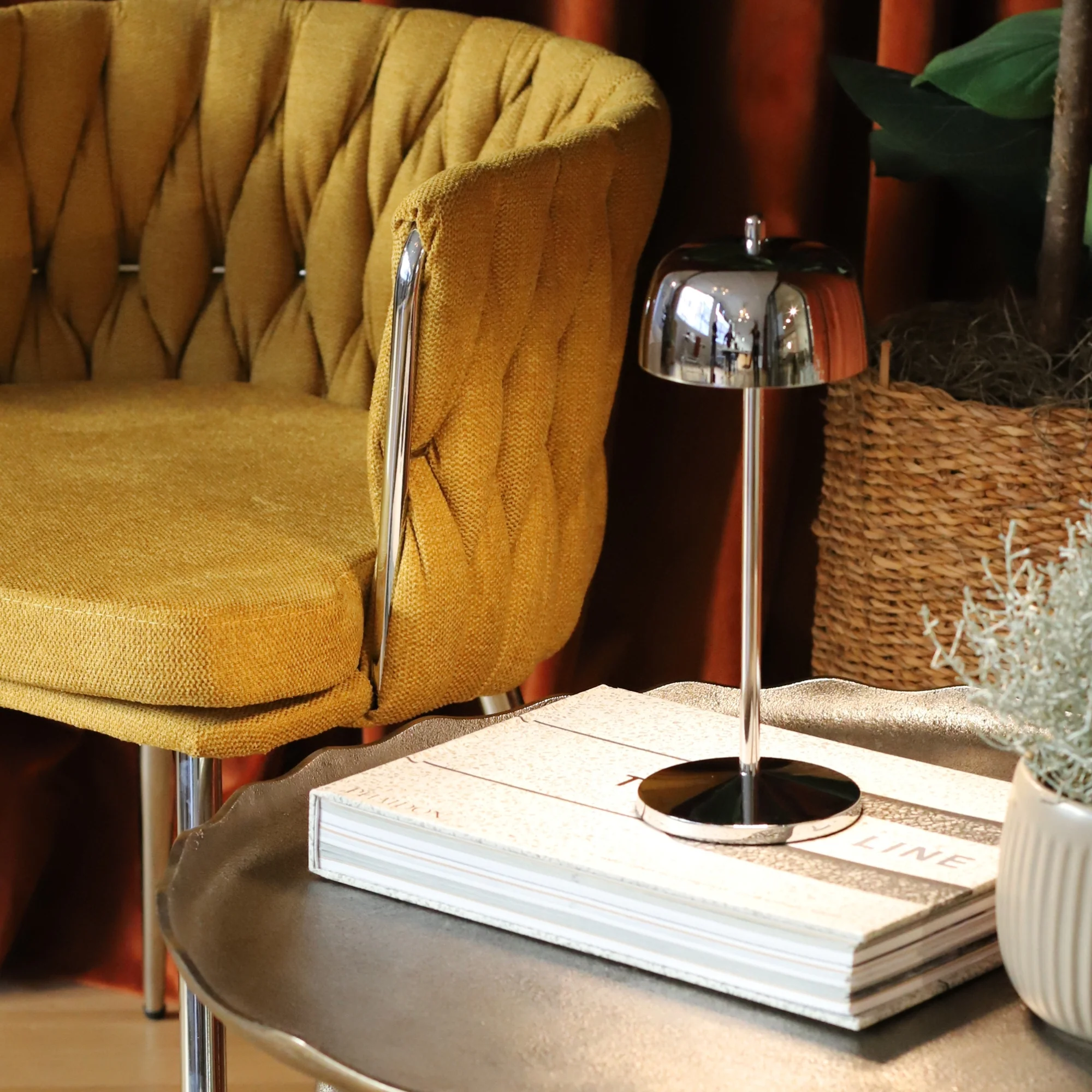 The polished chrome Theta Pro Cordless Lamp from Zafferano on a table next to a lounge chair in a hotel lobby.