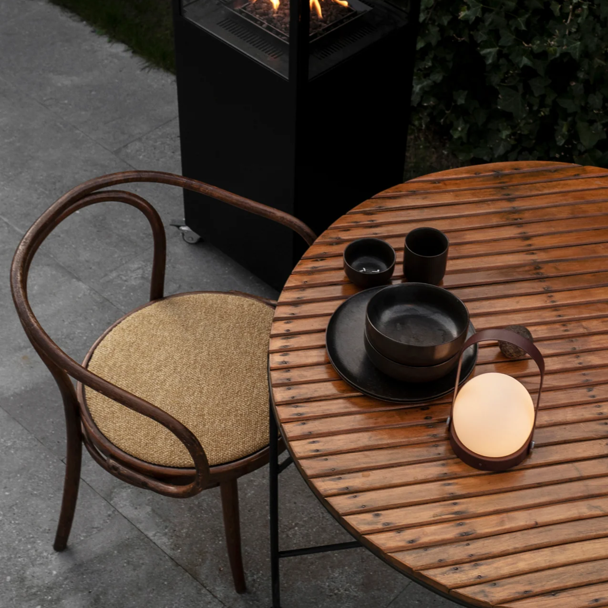 The burned red Carrie Portable Table Lamp from Audo Copenhagen on an outdoor dining table with plates, bowls and cups.