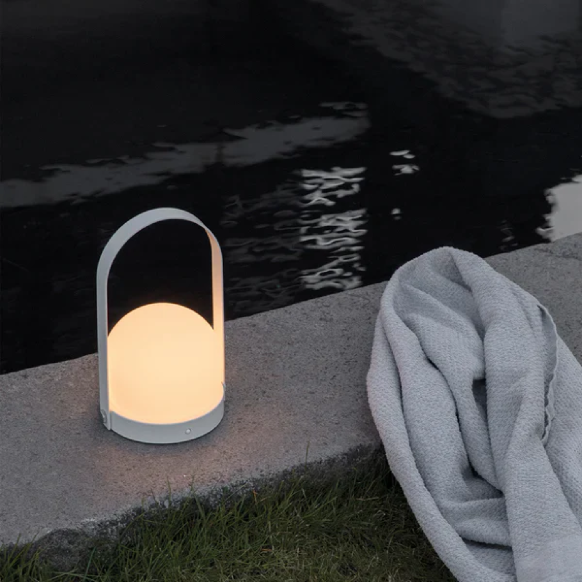 The white Carrie Portable Table Lamp from Audo Copenhagen beside a pool with a towel.