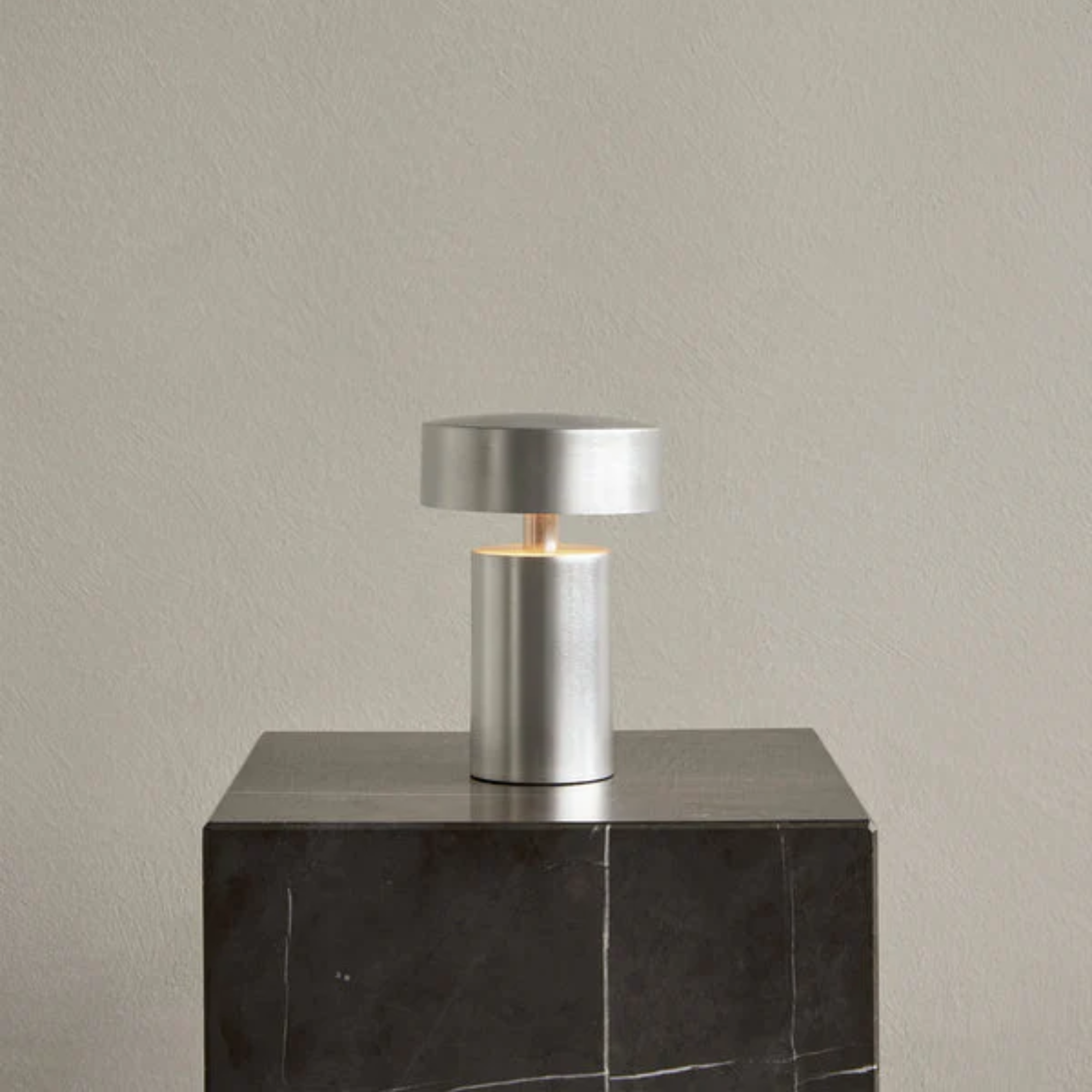 The brushed aluminum Column Portable Table Lamp from Audo Copenhagen in a living room on top of a marble side table plinth.