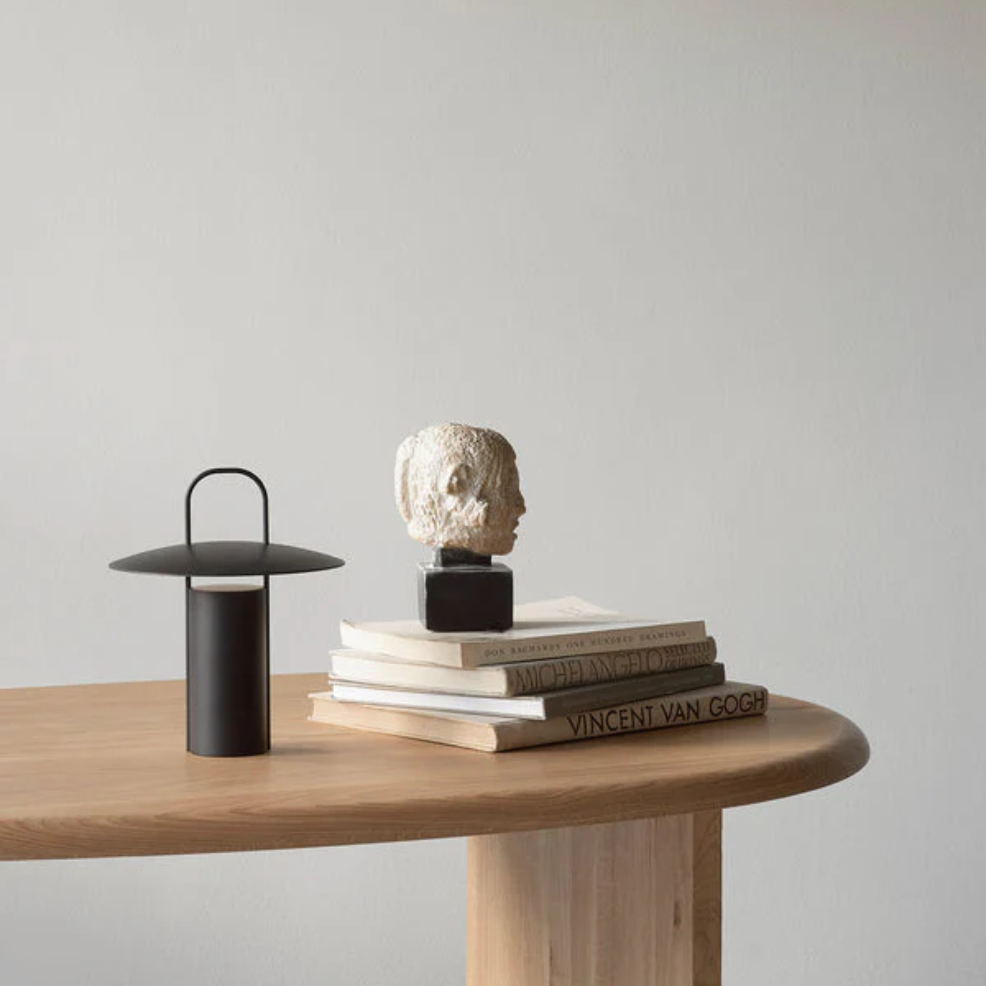 The black Ray Portable Table Lamp from Audo Copenhagen in a home office on a wood table beside a stack of books and a sculpture.