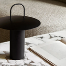 The black Ray Portable Table Lamp from Audo Copenhagen in a living room on a marble coffee table beside a seat.