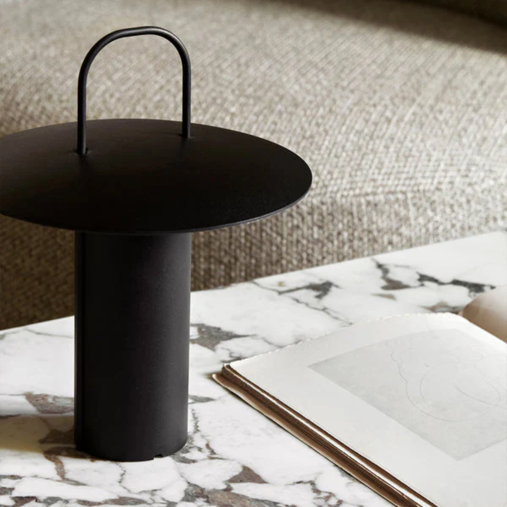 The black Ray Portable Table Lamp from Audo Copenhagen in a living room on a marble coffee table beside a seat.