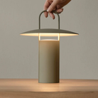 The dusty matte green Ray Portable Table Lamp from Audo Copenhagen in a lounge being placed onto a table.
