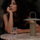 The dusty matte green Ray Portable Table Lamp from Audo Copenhagen on a dining table outdoors with a woman sitting at the table.
