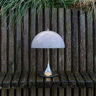 The grey opal Panthella 250 Portable Lamp from Louis Poulsen in a garden on a wooden bench in front of some plants.