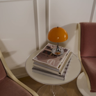 The 9.5 inch orange Panthella Portable Lamp: The Originals from Louis Poulsen on top of magazine in a living space, beside two chairs.