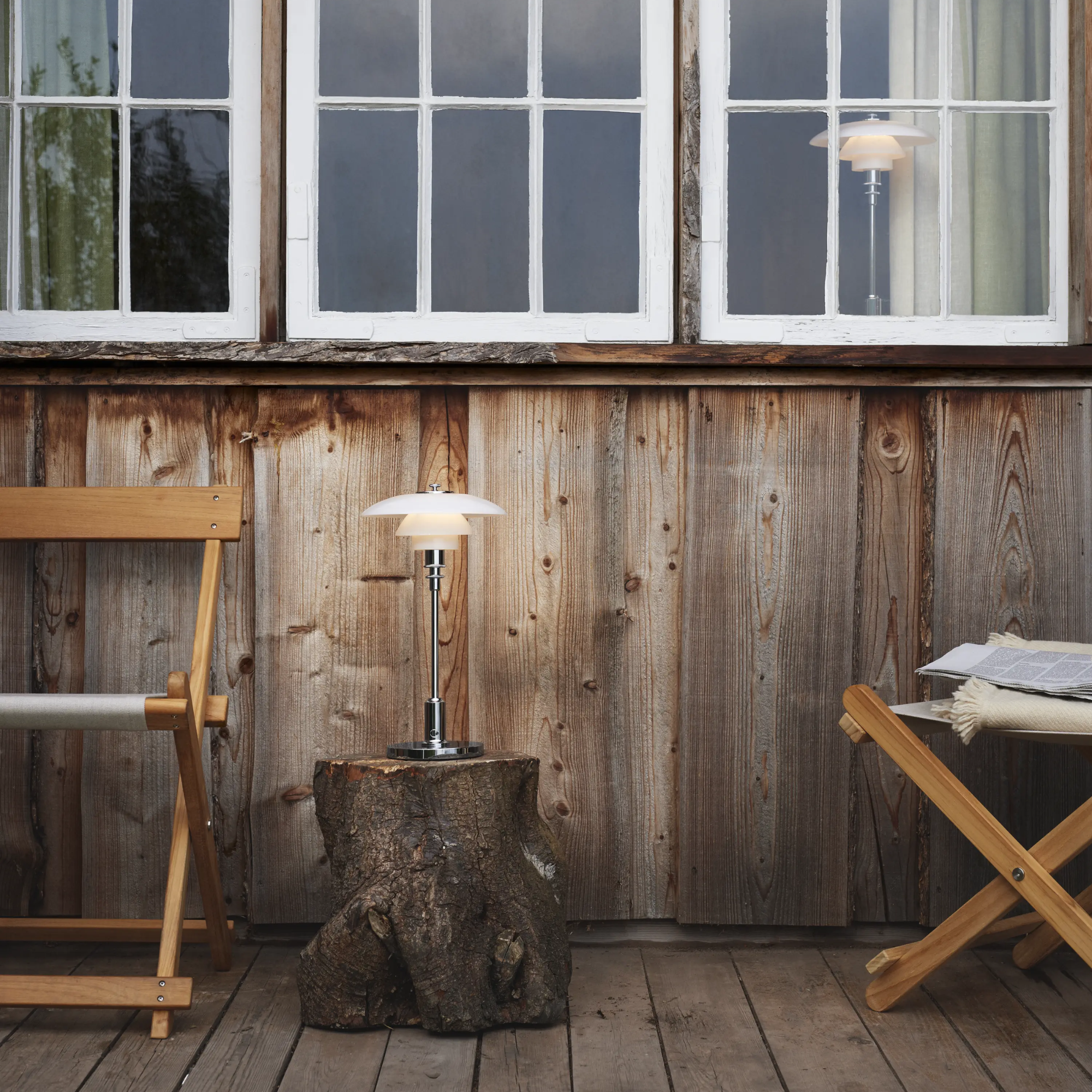 The chrome PH 2/1 Portable Lamp from Louis Poulsen on top of a log side table on an outdoor porch beside a chair and in the window.