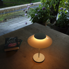 The opal white and white plate PH 80 Portable Lamp from Louis Poulsen on top of a wooden table overlooking a garden.