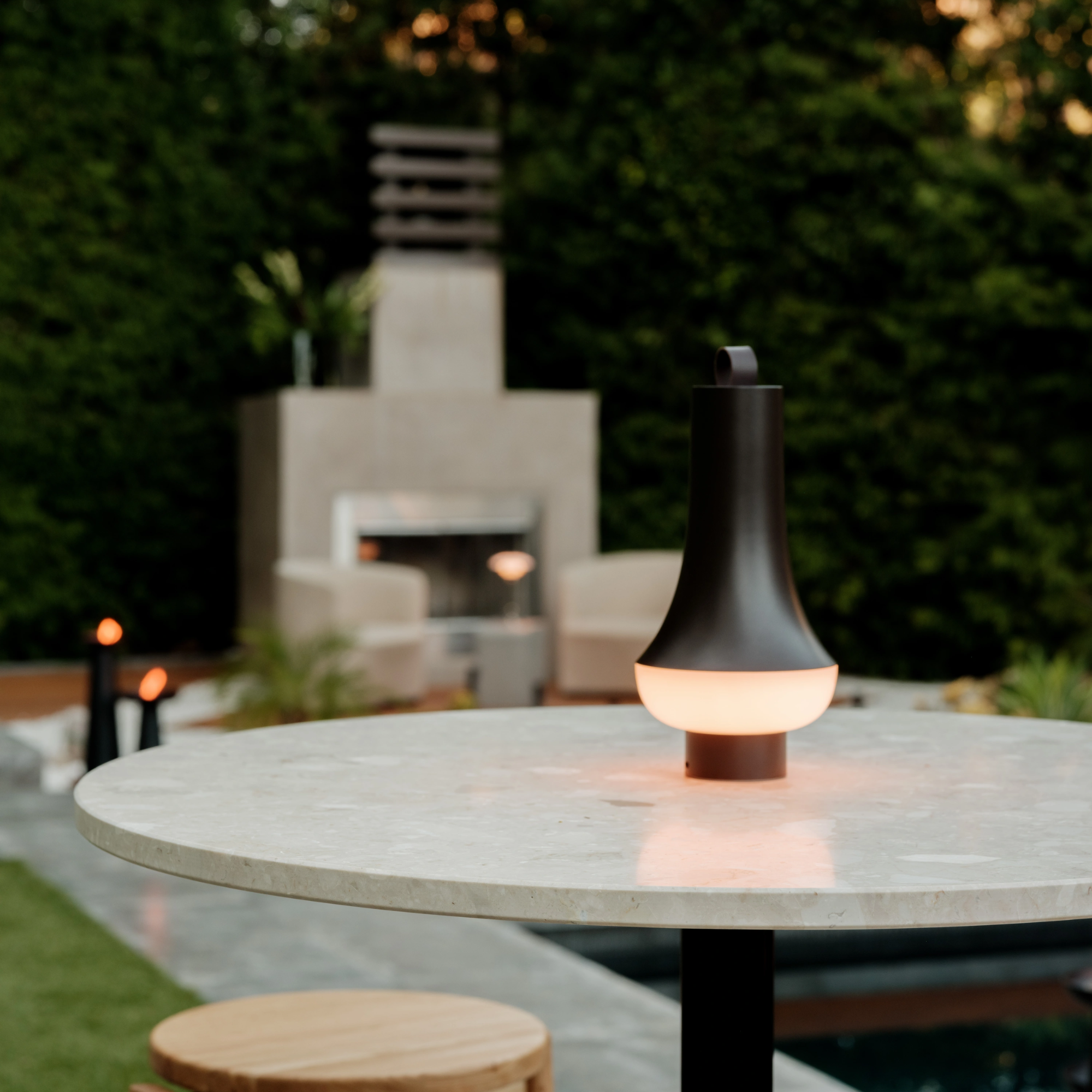 The dark brown Tomoshi Portable Lamp from Louis Poulsen on an outdoor marble dining table in a garden beside a pool.