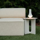 The white Tomoshi Portable Lamp from Louis Poulsen in an outdoor garden on top of a side table next to a sofa.