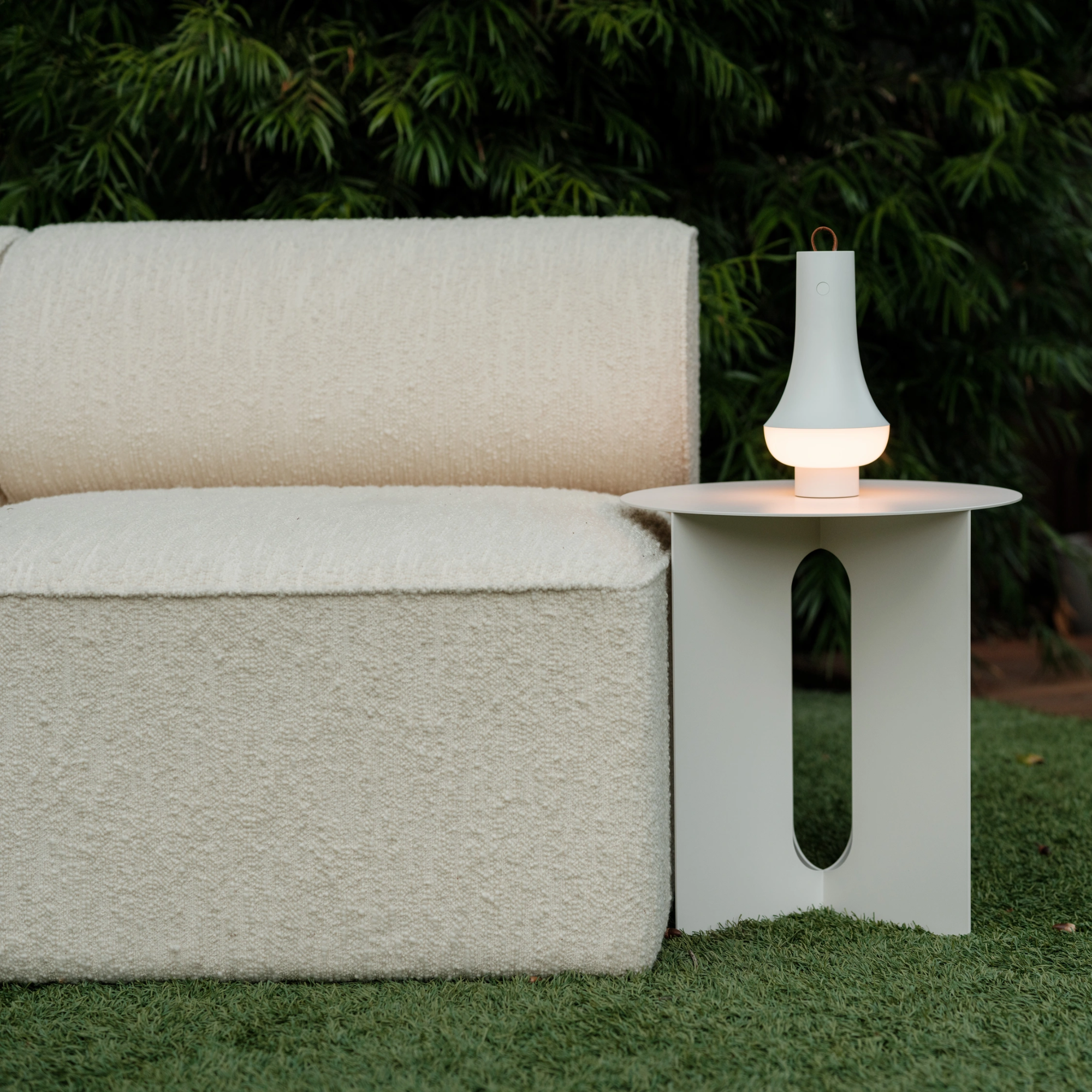 The white Tomoshi Portable Lamp from Louis Poulsen in an outdoor garden on top of a side table next to a sofa.
