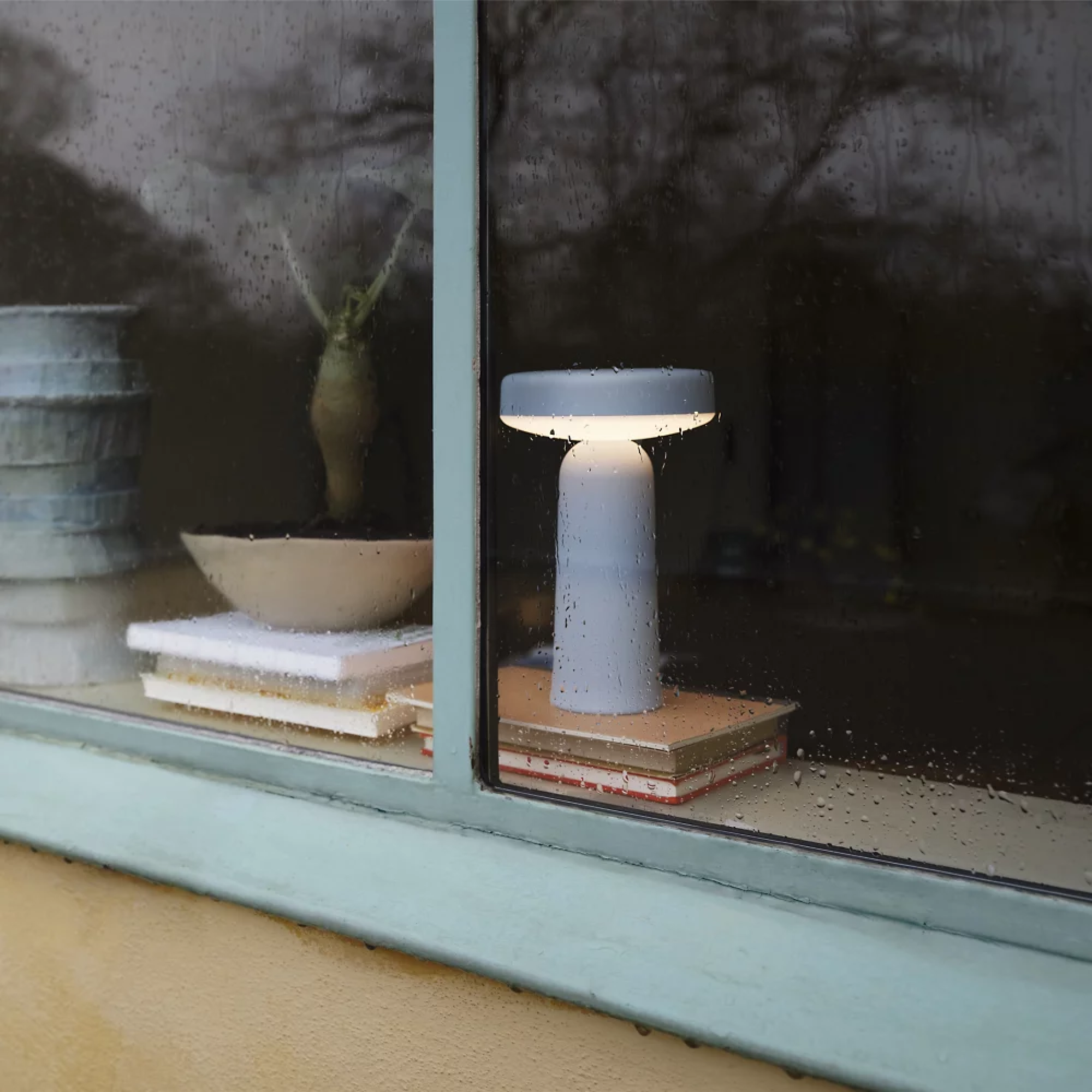 The light blue Ease Cordless Lamp from Muuto in a living room through a rainy window.