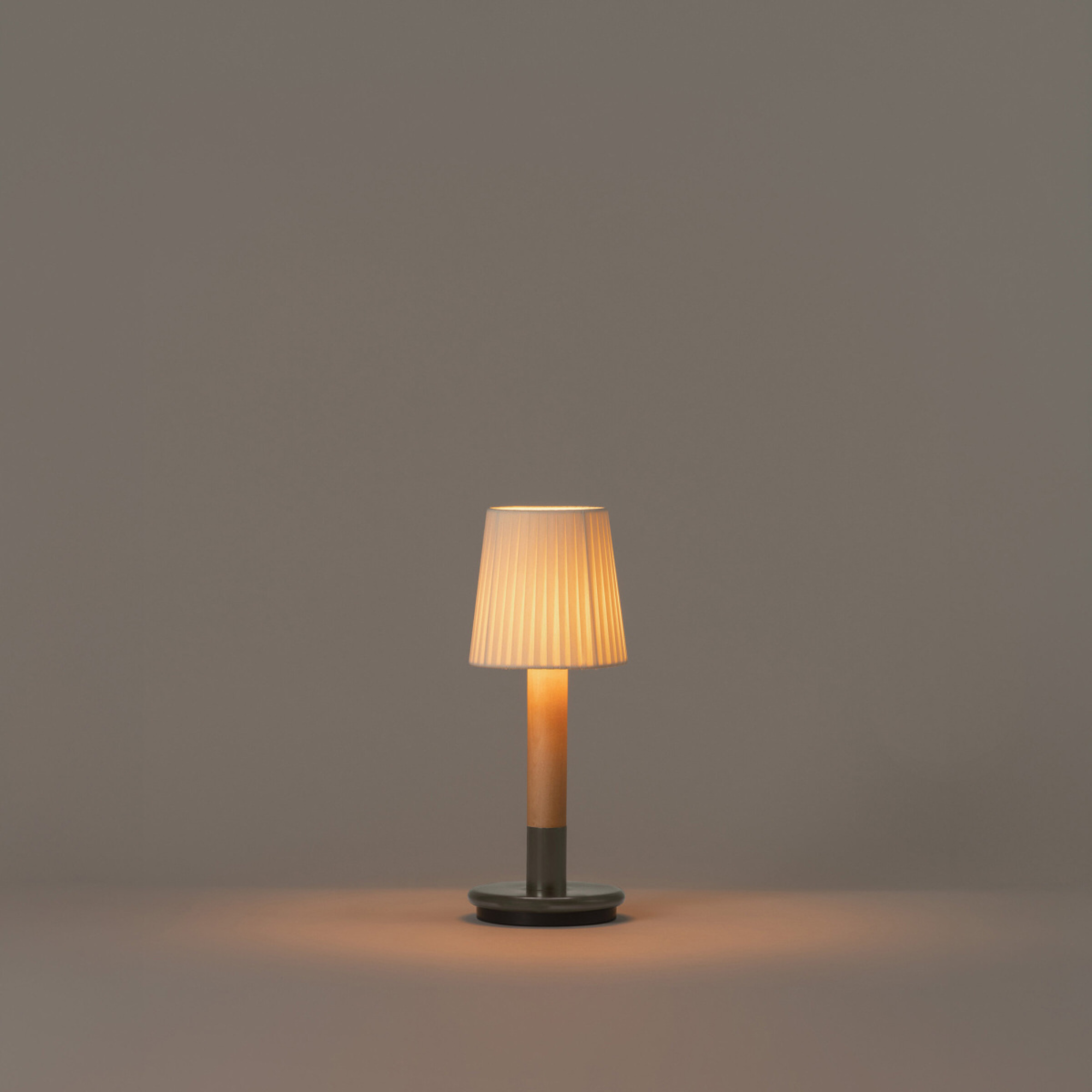 The Básica Mínima Batería Cordless Lamp from Santa & Cole with the natural ribbon shade turned on in a studio.
