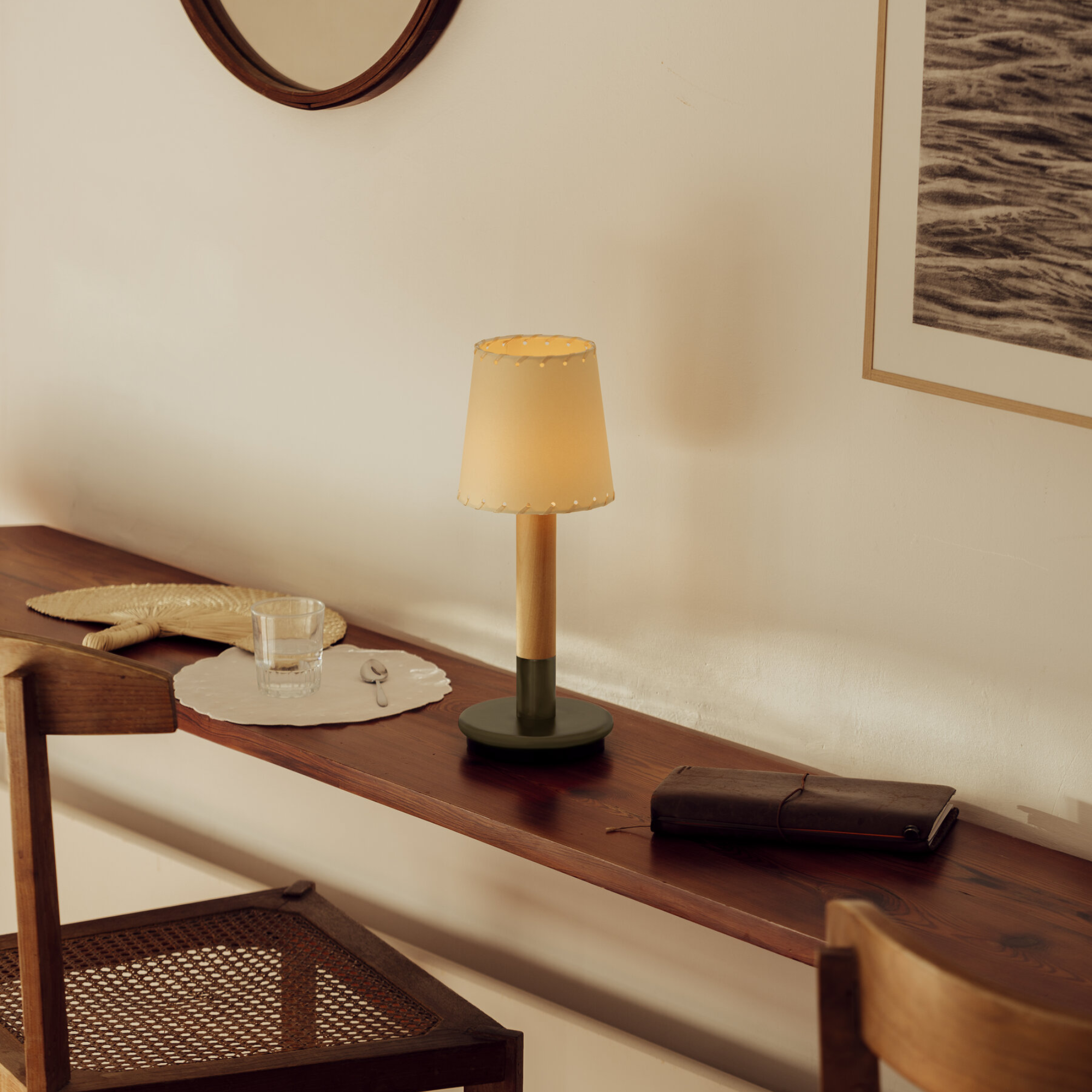 The Básica Mínima Batería Cordless Lamp from Santa & Cole with the stitched beige parchment shade in a living room on a wooden surface.