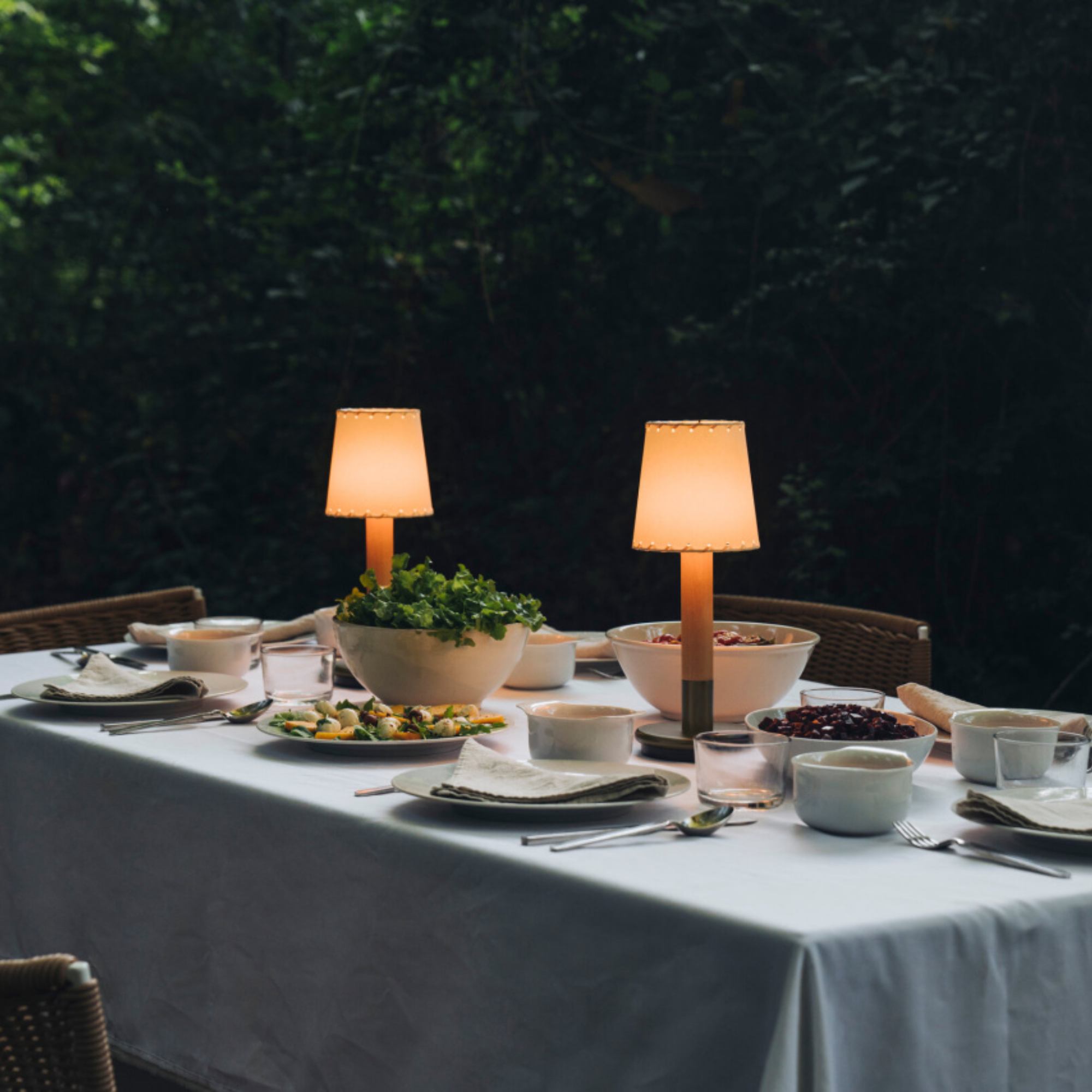The Básica Mínima Batería Cordless Lamp from Santa & Cole with the stitched beige parchment shade on an outdoor dining table.