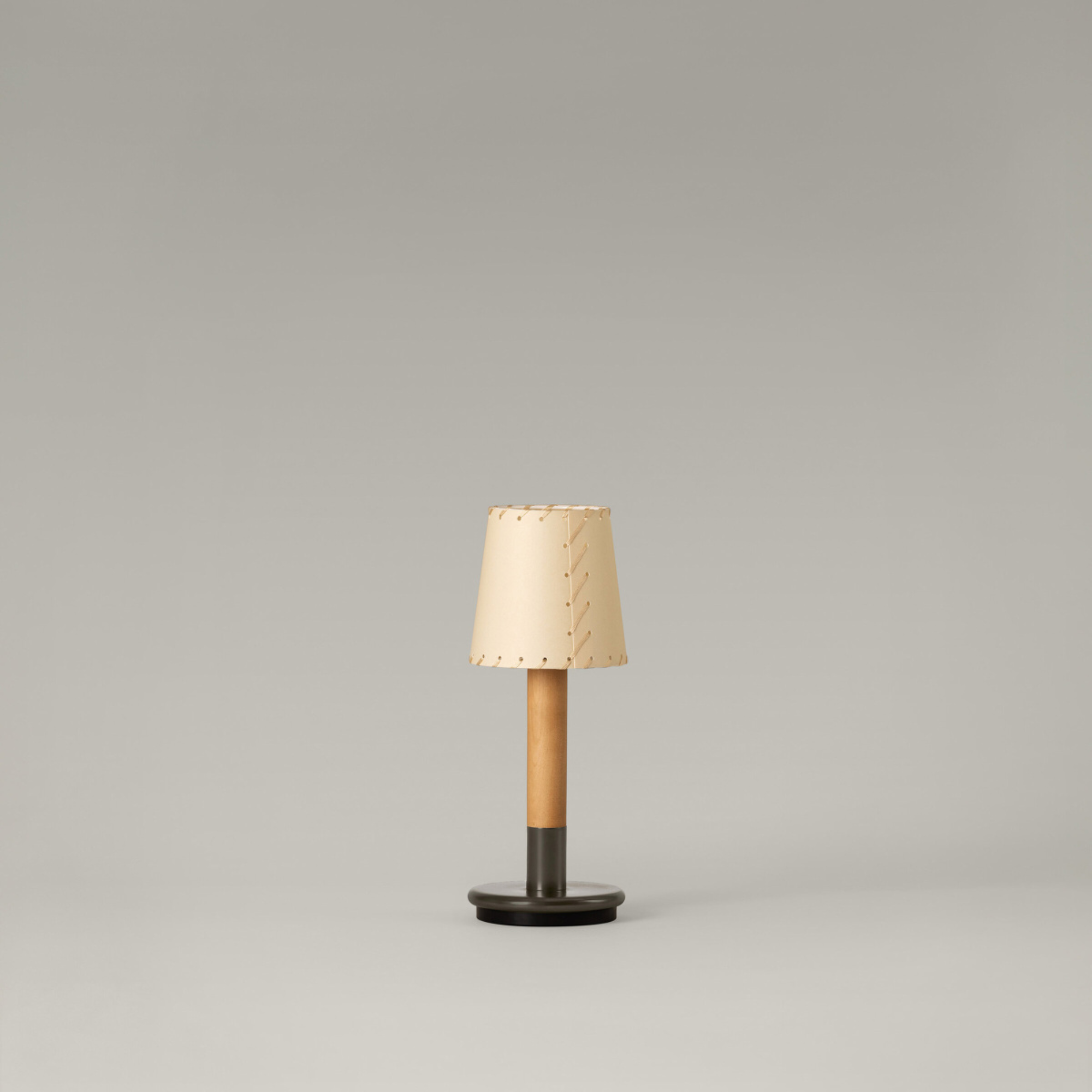 The Básica Mínima Batería Cordless Lamp from Santa & Cole with the stitched beige parchment shade turned off in a studio.