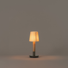 The Básica Mínima Batería Cordless Lamp from Santa & Cole with the stitched beige parchment shade turned on in a studio.