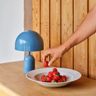 The baby blue Bell Cordless Lamp from Tom Dixon in a living space on a wooden counter.