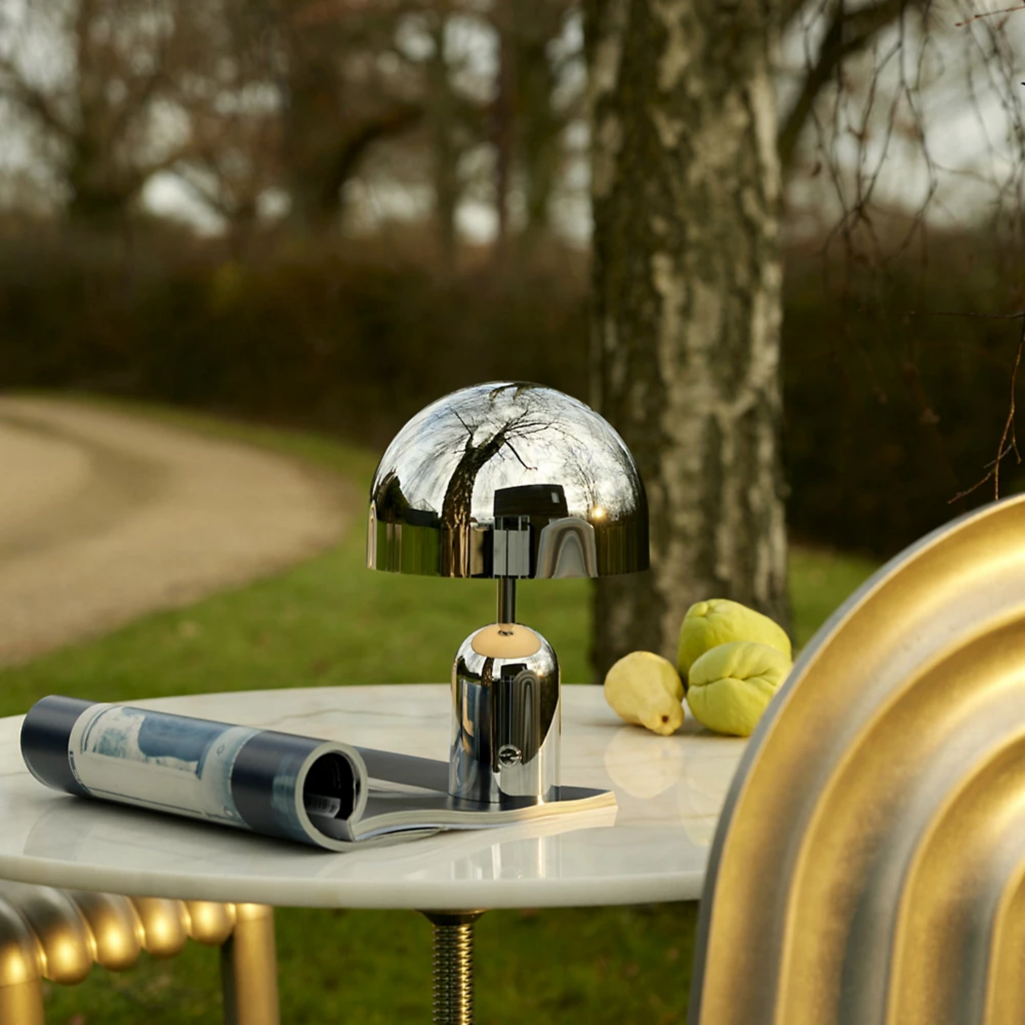 The silver Bell Cordless Lamp from Tom Dixon on a cafe table outdoors overlooking a garden path.