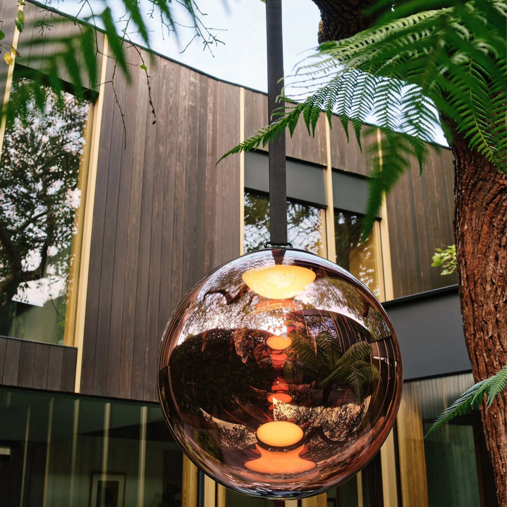 The copper Melt Cordless Floor Lamp from Tom Dixon hanging on a tree in a garden using the included strap.