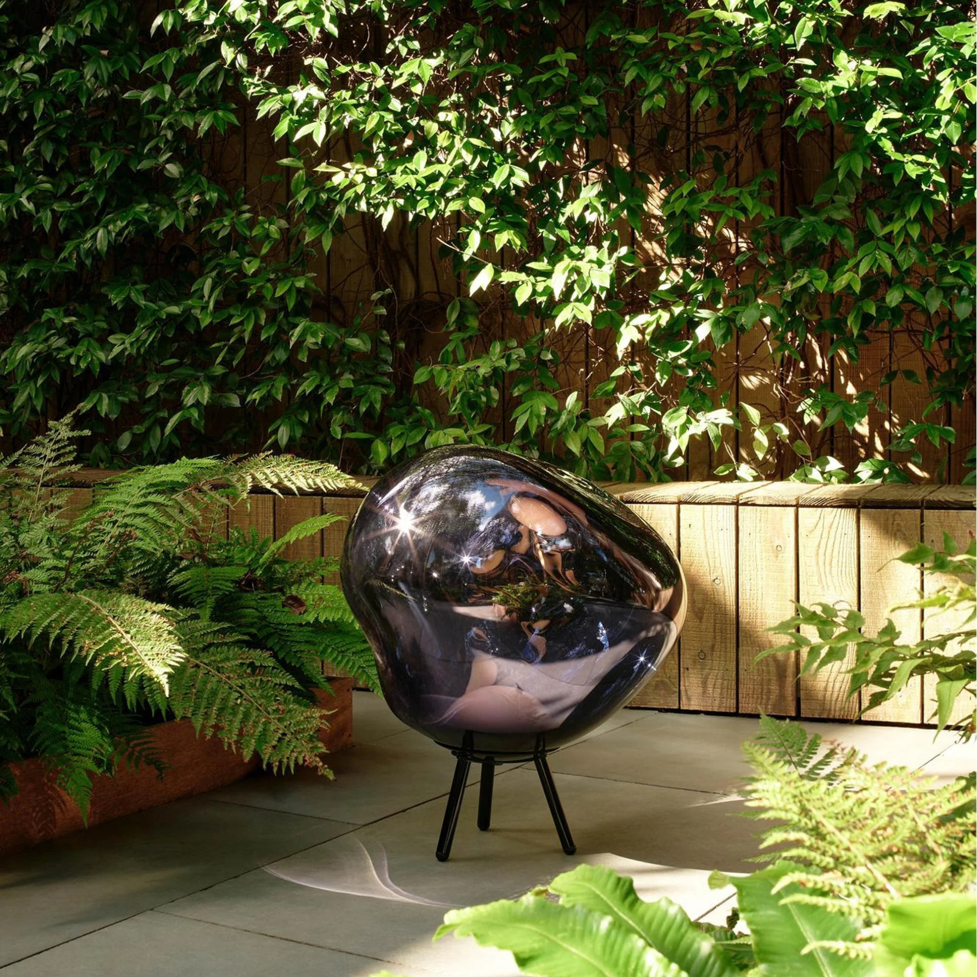 The smoke Melt Cordless Floor Lamp from Tom Dixon in a courtyard beside various trees and ferns.
