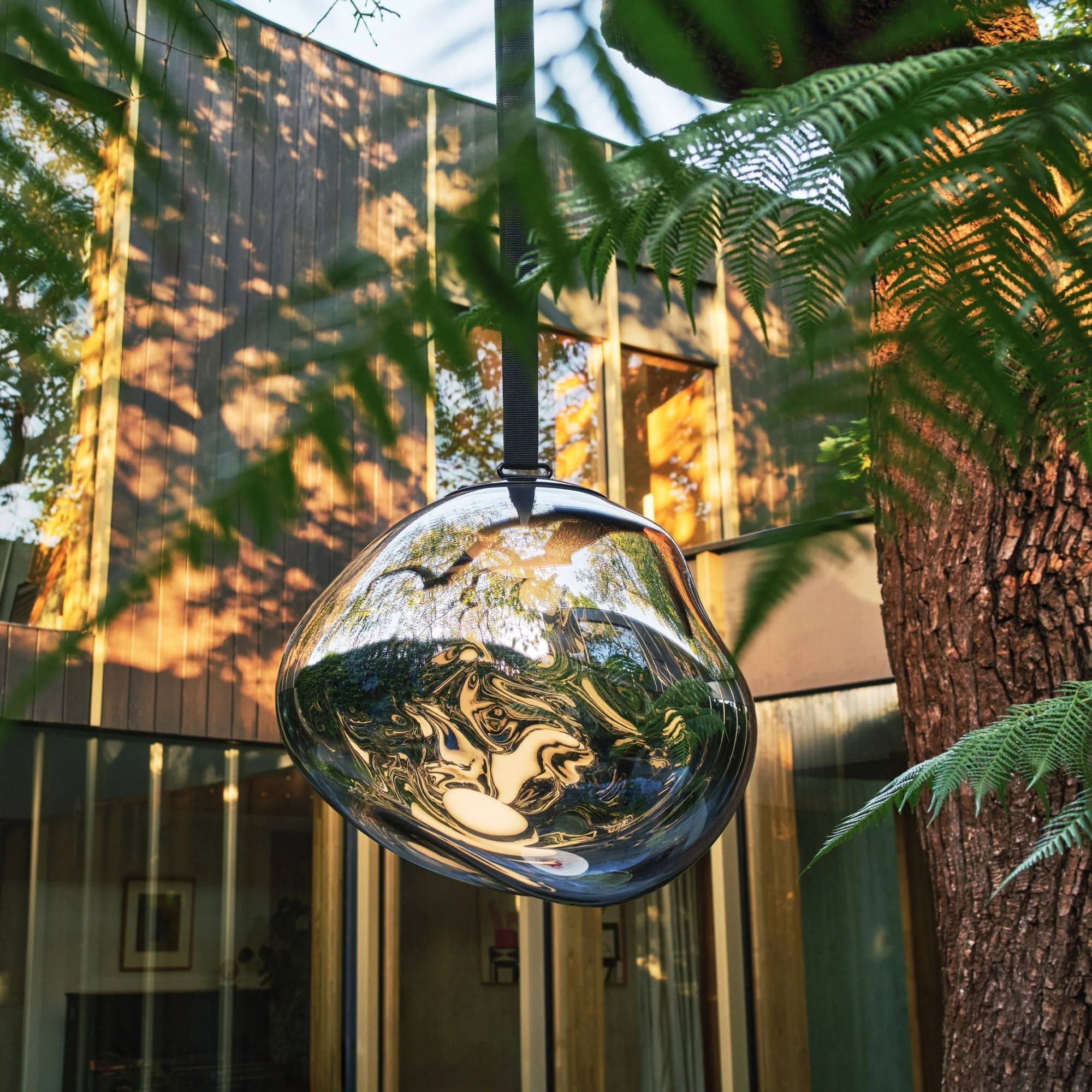 The smoke Melt Cordless Floor Lamp from Tom Dixon hanging from a tree in a garden.