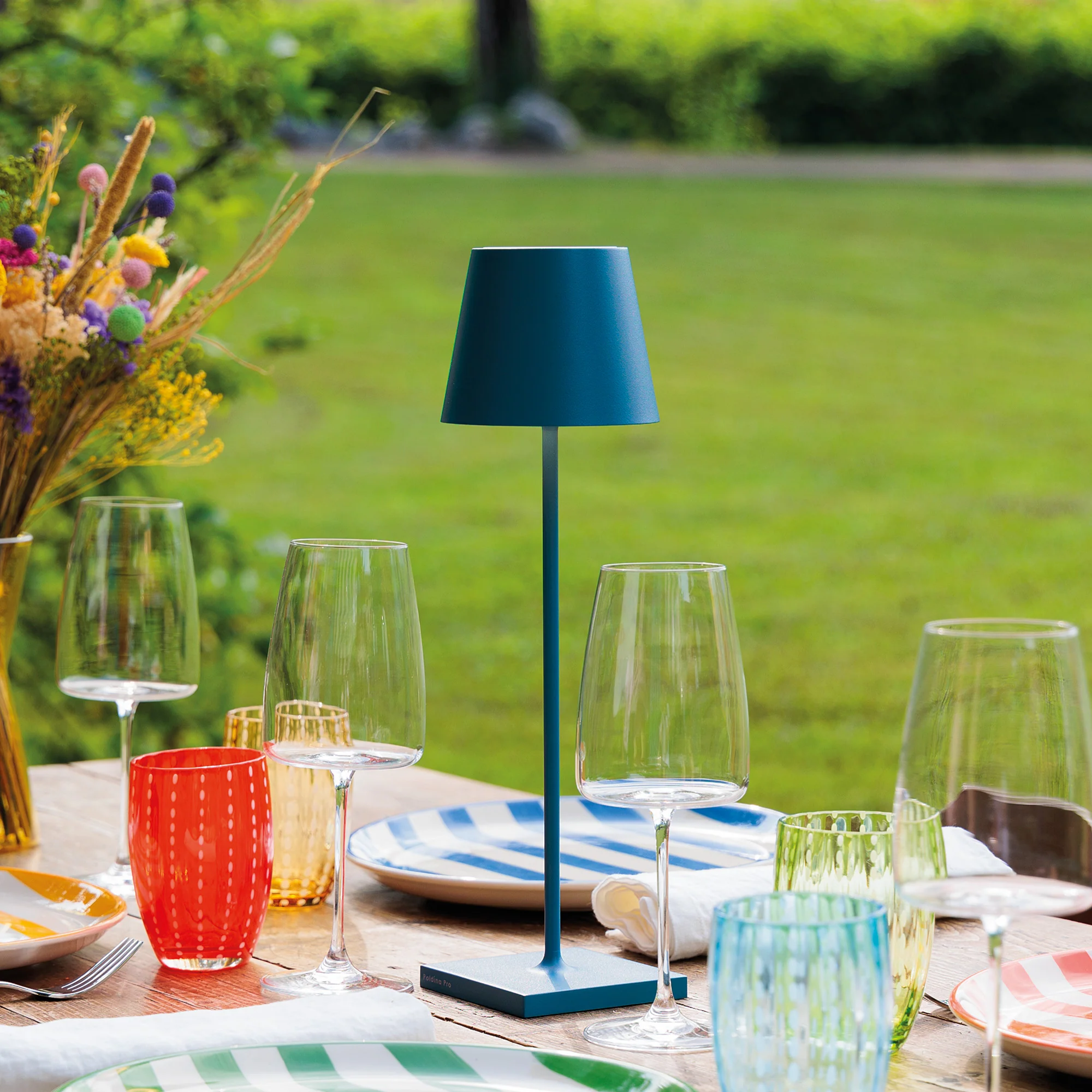 The avio blue Poldina Pro from Zafferano in a garden on a dining table with glasses and dishes.