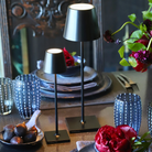 The black Poldina Pro Micro Cordless Lamp from Zafferano on a dining table with the Poldina Pro and various pllates, bowls and glasses.