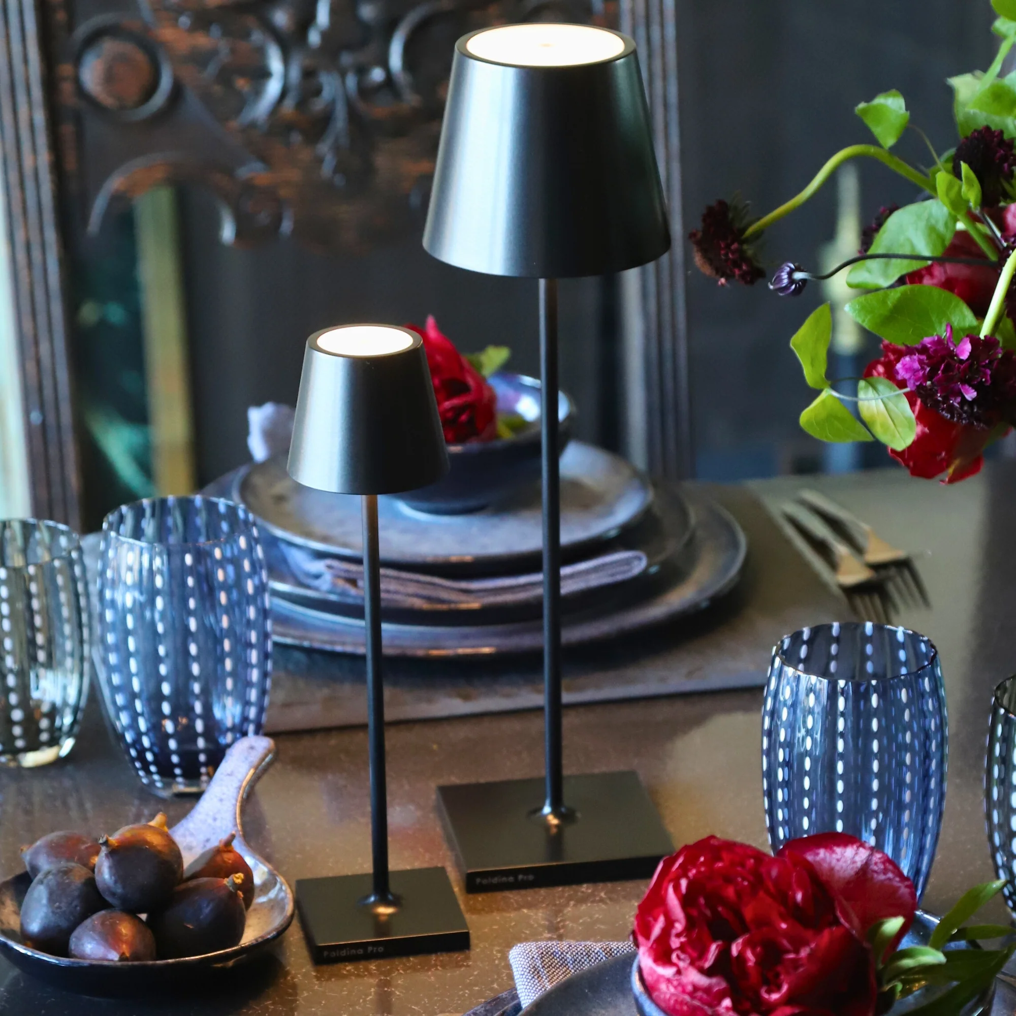 The black Poldina Pro Micro Cordless Lamp from Zafferano on a dining table with the Poldina Pro and various pllates, bowls and glasses.