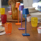 The capri blue Poldina Pro Micro Cordless Lamp from Zafferano on a table at an outdoor dinner party with various other colors.