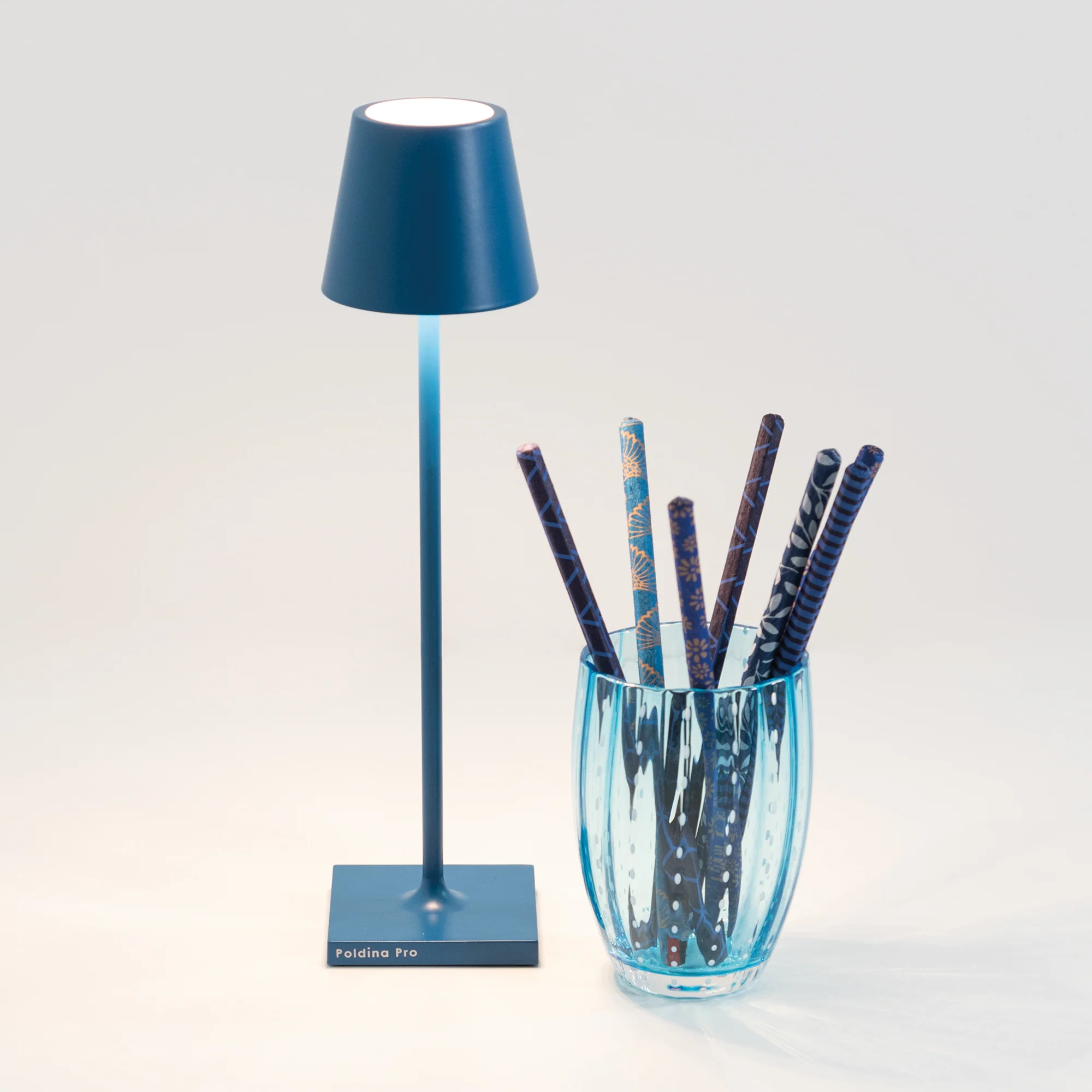 The capri blue Poldina Pro Micro Cordless Lamp from Zafferano in a studio beside a cup.