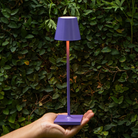 The lilac Poldina Pro Micro Cordless Lamp from Zafferano being held in front of a bush in a garden.