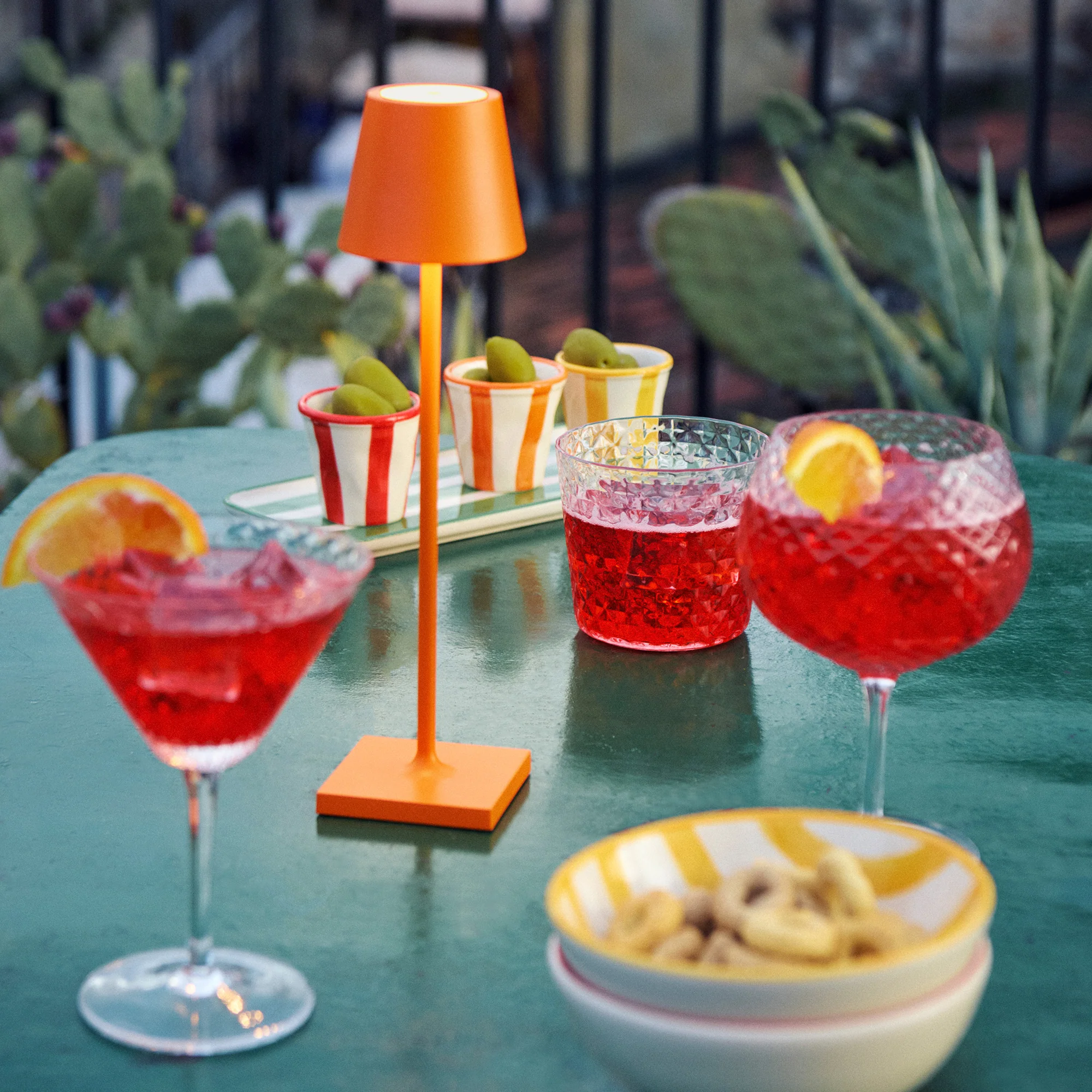 The orange Poldina Pro Micro Cordless Lamp from Zafferano on a dining table outside with glasses.