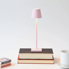 The pink Poldina Pro Micro Cordless Lamp from Zafferano in a library studio on top of a stack of books.