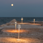 The white Poldina Pro Micro Cordless Lamp from Zafferano illuminating an outdoor path beside a lake.